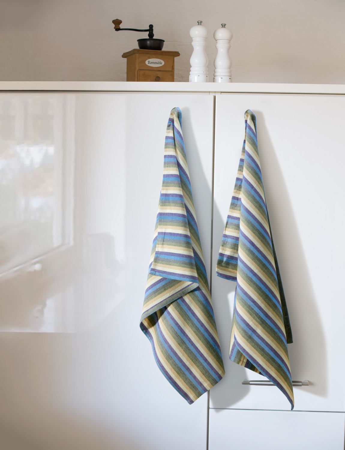 Zwei Baumwoll-Geschirrtücher mit diagonal verlaufenden Streifen in Blau, Grün, Beige und Weiß hängen an weißen Küchenschranktüren, darüber stehen eine alte Holz-Kaffeemühle und zwei weiße Gewürzmühlen.