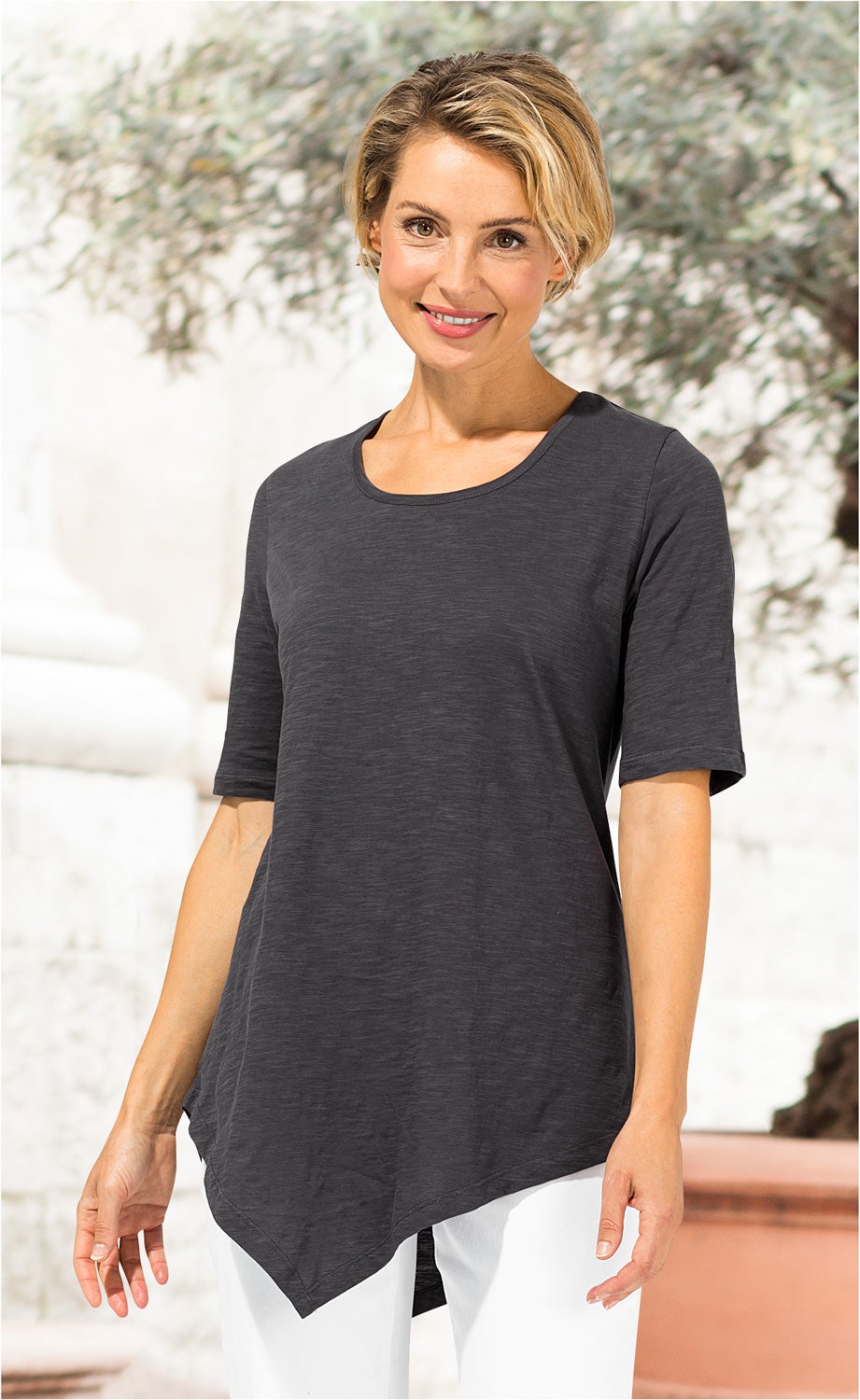Frau trägt dunkelgraues Baumwoll-Shirt mit asymmetrischem Saum und halblangen Ärmeln, kombiniert mit weißen Hosen, vor hellem, unscharfem Hintergrund mit Pflanzen und Mauerwerk.