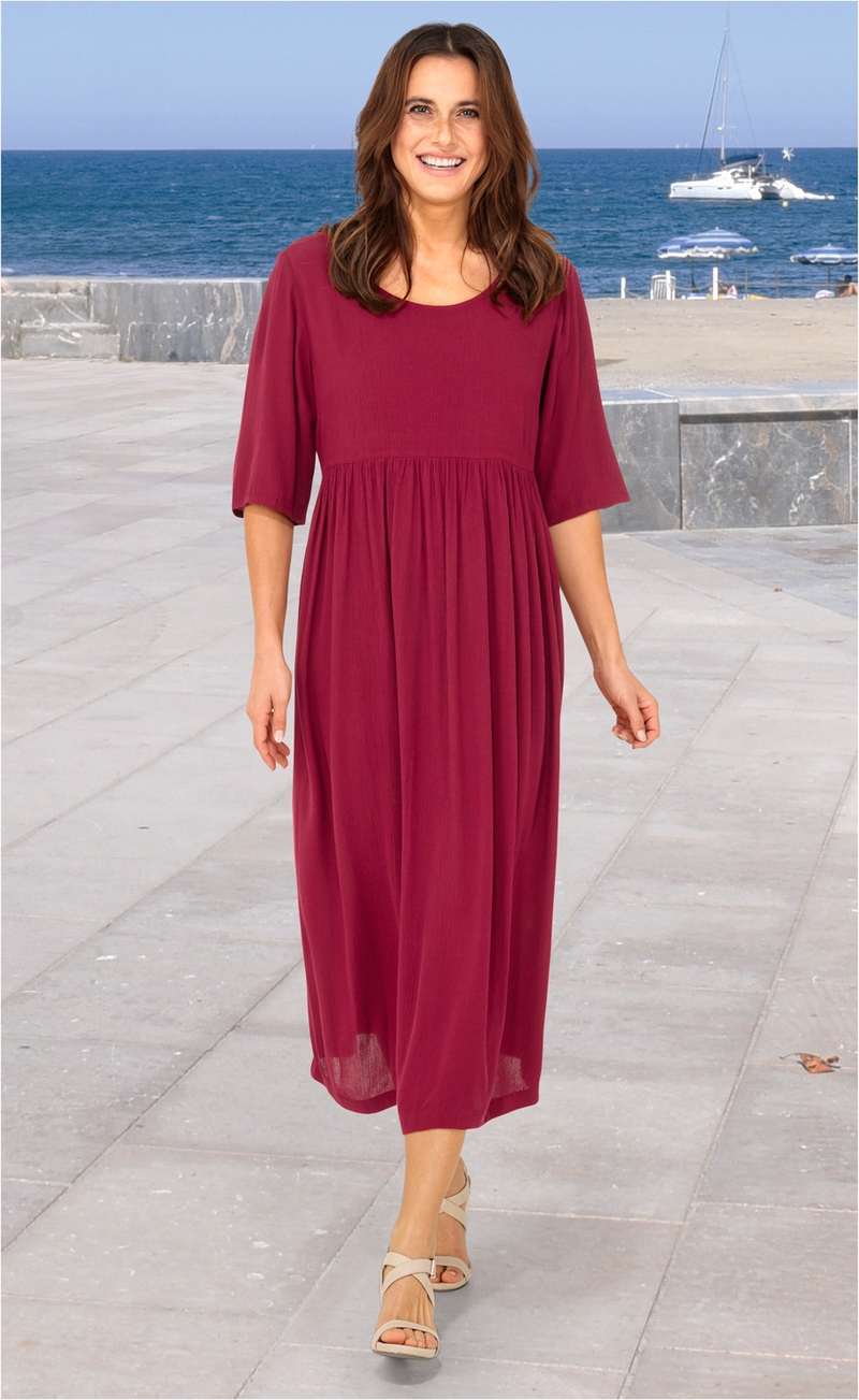 Frau läuft am Strandpromenade, trägt langes, locker fallendes, dunkelrotes Viskose-Kleid mit halblangen Ärmeln und Sandalen, im Hintergrund Meer, Segelboot und Strand mit Sonnenschirmen sichtbar