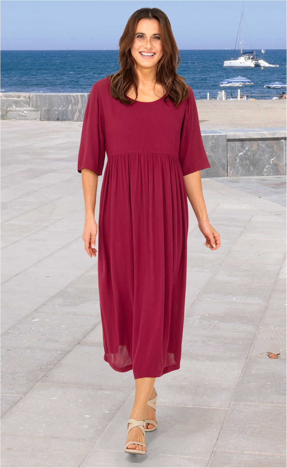 Frau läuft am Strandpromenade, trägt langes, locker fallendes, dunkelrotes Viskose-Kleid mit halblangen Ärmeln und Sandalen, im Hintergrund Meer, Segelboot und Strand mit Sonnenschirmen sichtbar