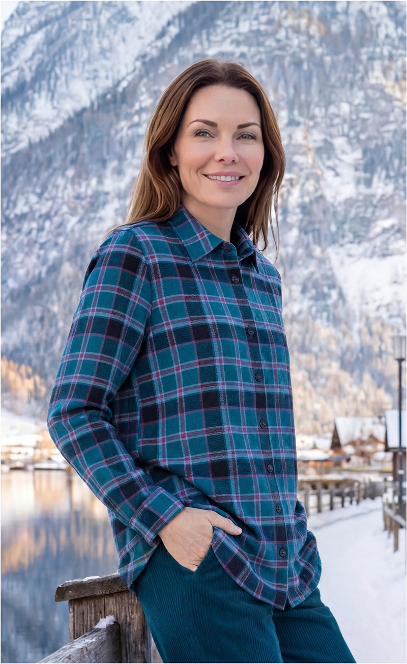 Frau mit braunen Haaren trägt kariertes Hemd und blaue Hose, steht am Holzgeländer vor winterlicher Berglandschaft mit See und verschneiten Häusern im Hintergrund, lächelnd, Hand in Hosentasche