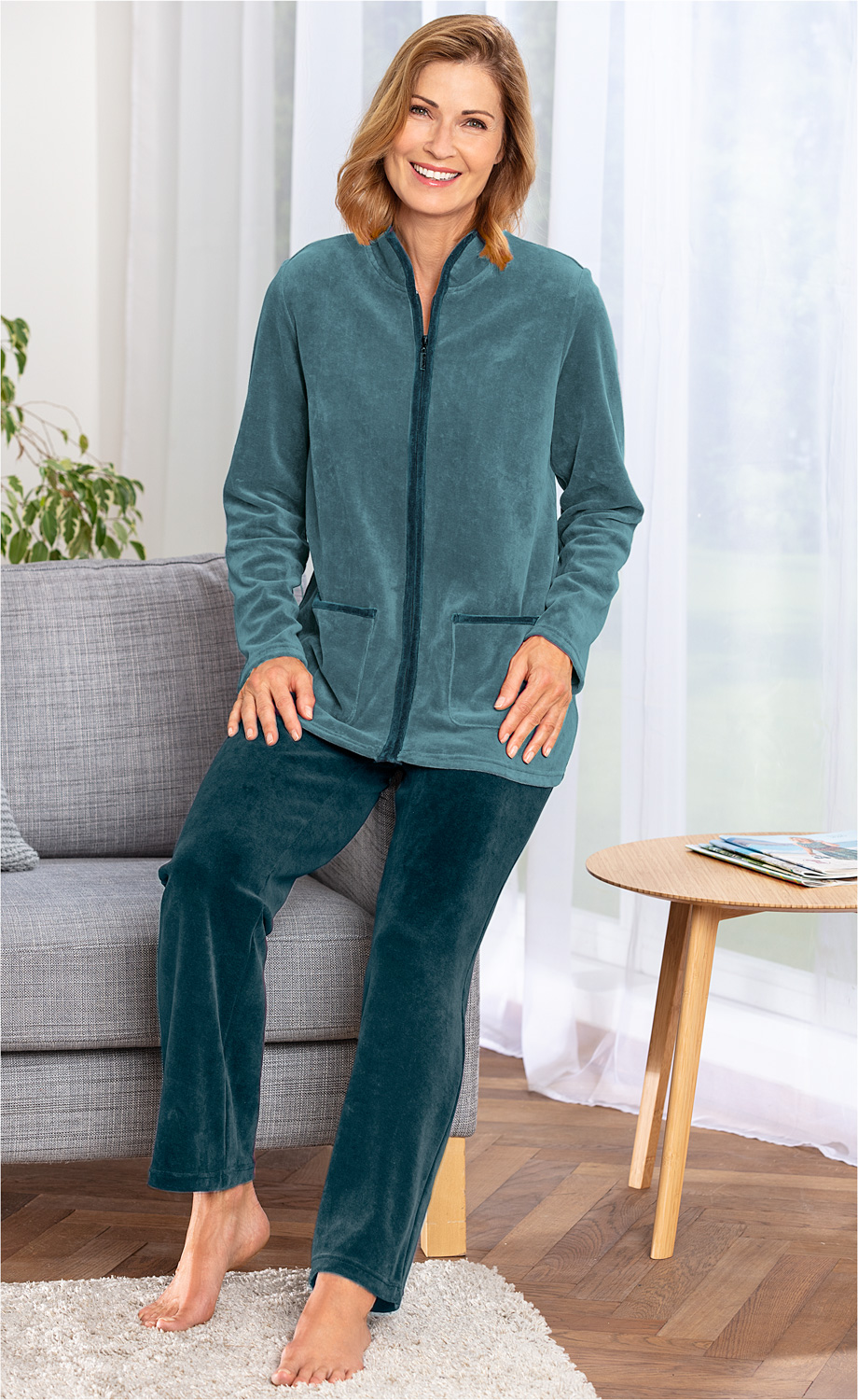 Frau sitzt barfuß auf grauem Sofa, trägt blauen weichen Baumwoll-Hausanzug mit Reißverschluss und Taschen, heller Raum mit Holzboden, weißer Gardine und rundem Holztisch mit Zeitschriften im Hintergrund.
