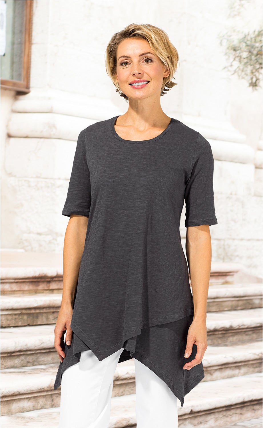 Frau trägt dunkelgraues, asymmetrisch geschnittenes Baumwoll-Shirt mit kurzen Ärmeln und Rundhalsausschnitt, kombiniert mit weißen Hosen, vor hellen Marmortreppen stehend
