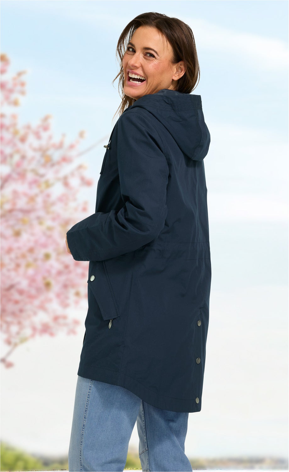 Frau mit dunkler Baumwoll-Jacke mit Kapuze, seitlich stehend, lachend, vor unscharfem Frühlingshintergrund mit blühendem Baum und blauem Himmel, trägt helle Jeans