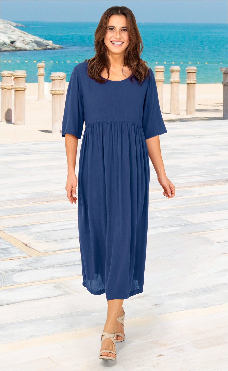 Frau am Strand trägt ein knielanges, blaues Viskose-Kleid mit halblangen Ärmeln und beigen Sandaletten, sonniger Tag mit Meer und Felsen im Hintergrund