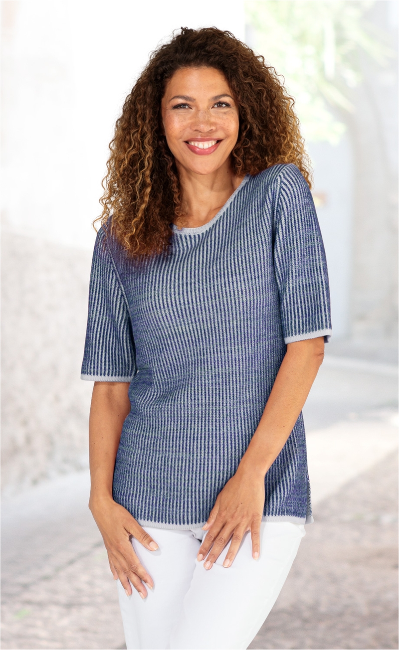 Frau mit lockigem Haar, lächelnd, trägt blau-grau gestreiften Baumwoll-Pullover mit halblangen Ärmeln und weiße Hose, stehend vor hellem, unscharfem Hintergrund