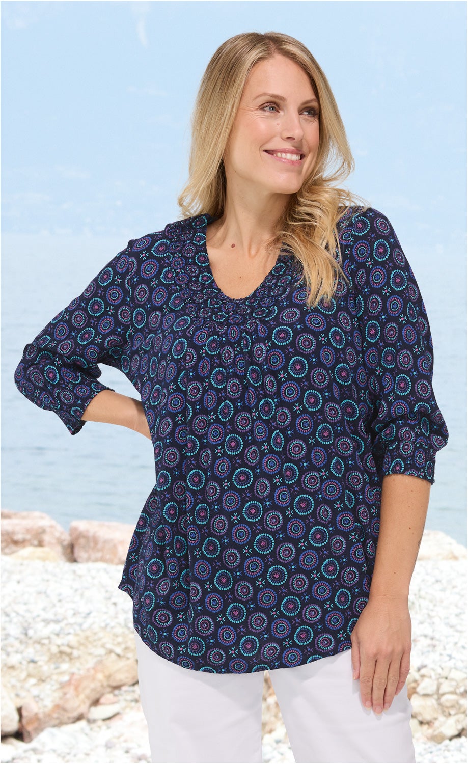 Frau trägt dunkelblaue Viskose-Bluse mit buntem Mandala-Muster, 3/4-Ärmeln und V-Ausschnitt, weiße Hose, steht am Kieselstrand mit Meer im Hintergrund, zeigt seitlichen Blick und lächelt.