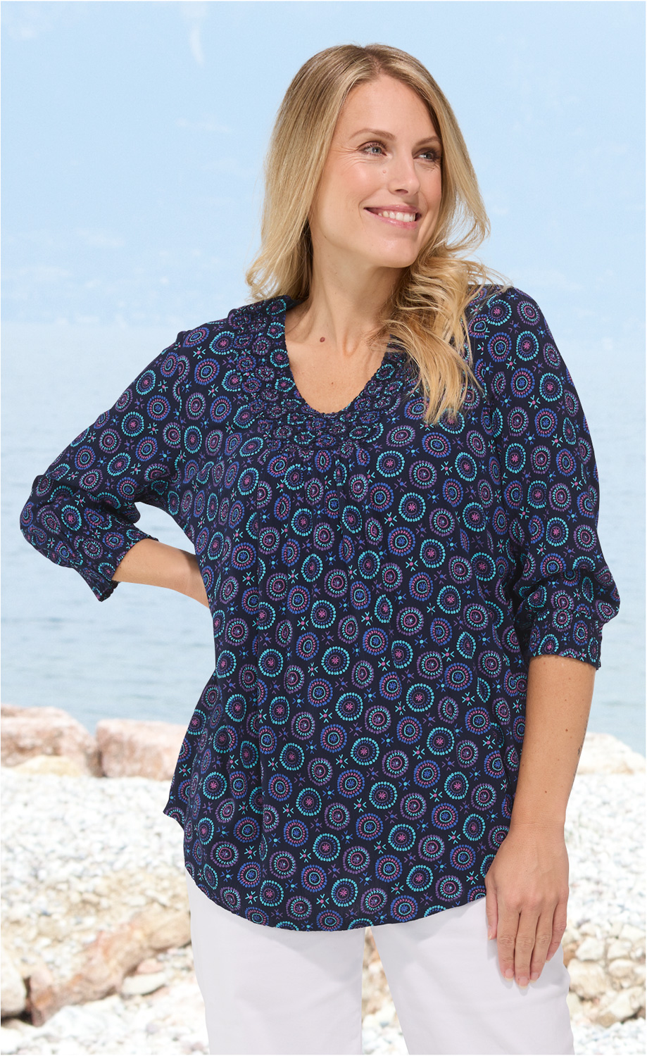 Frau trägt dunkelblaue Viskose-Bluse mit buntem Mandala-Muster, 3/4-Ärmeln und V-Ausschnitt, weiße Hose, steht am Kieselstrand mit Meer im Hintergrund, zeigt seitlichen Blick und lächelt.