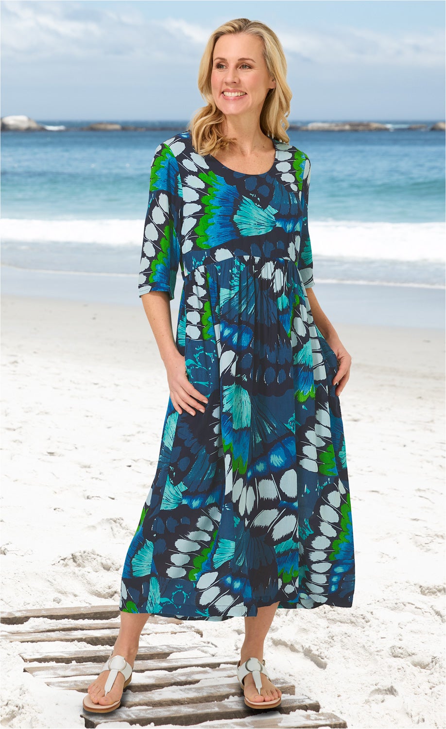 Frau am Strand trägt langes Viskose-Kleid mit blau-grünem Schmetterlingsmuster, halblangen Ärmeln und rundem Ausschnitt, weiße Sandalen, lächelnd, weißer Sand und Meer im Hintergrund