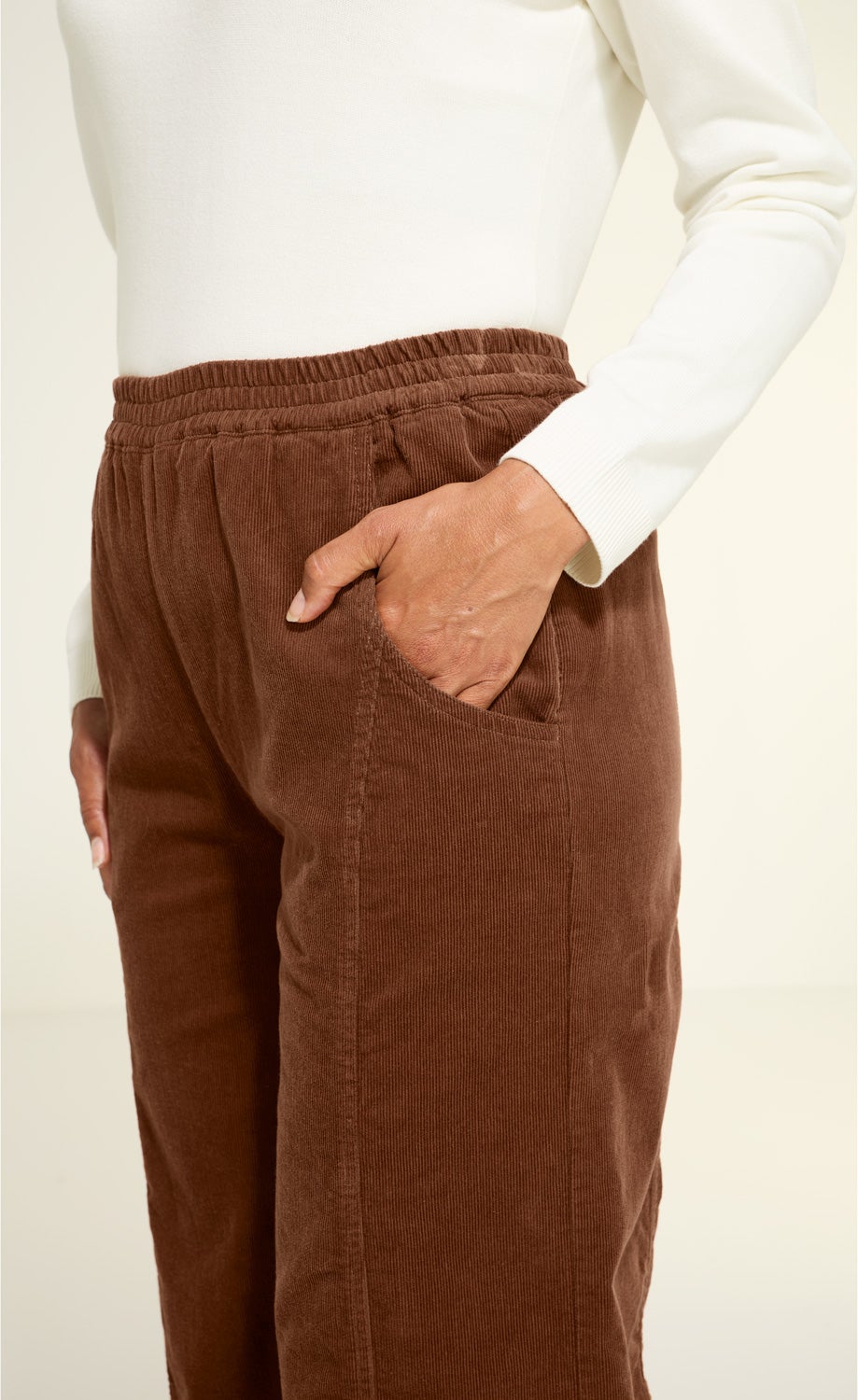 Braune Baumwoll-Hose mit elastischem Bund und aufgesetzten Taschen, Detailaufnahme von Hüfte und Hand in Tasche, Person trägt weißen Langarmpullover.