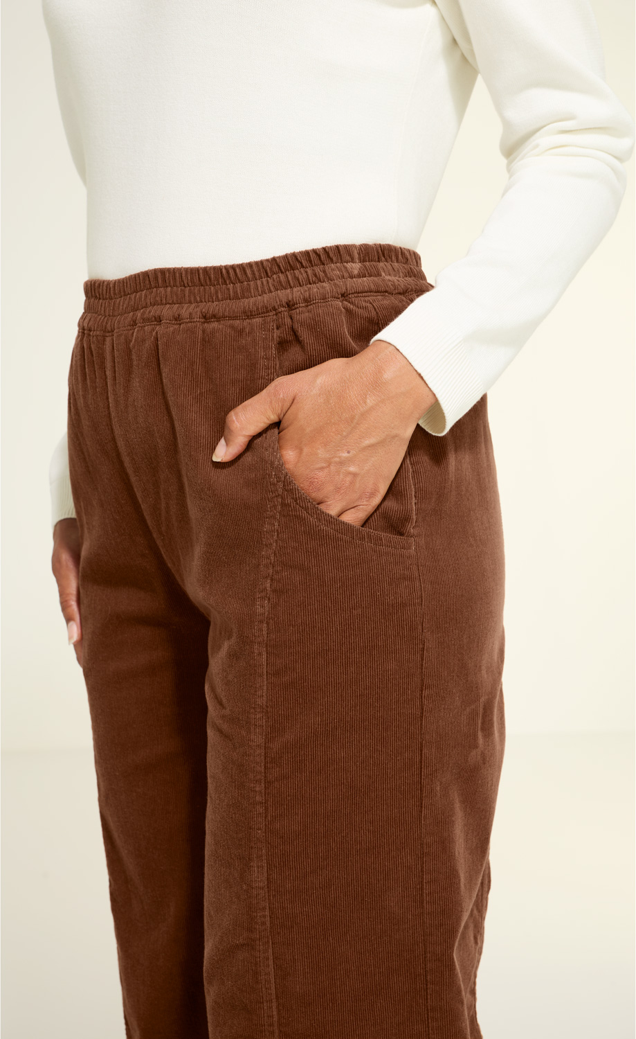 Braune Baumwoll-Hose mit elastischem Bund und aufgesetzten Taschen, Detailaufnahme von Hüfte und Hand in Tasche, Person trägt weißen Langarmpullover.