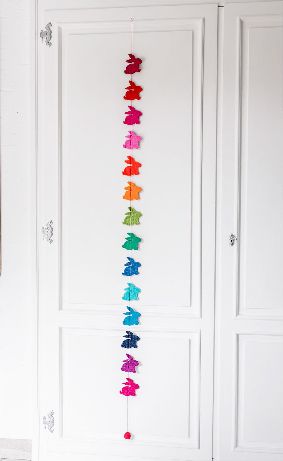 Bunte Girlande aus 12 filzartigen Hasen in Farben von Rot, Orange, Grün, Blau bis Pink, hängend an weißer Tür, unten Abschluss mit rotem Filzball.