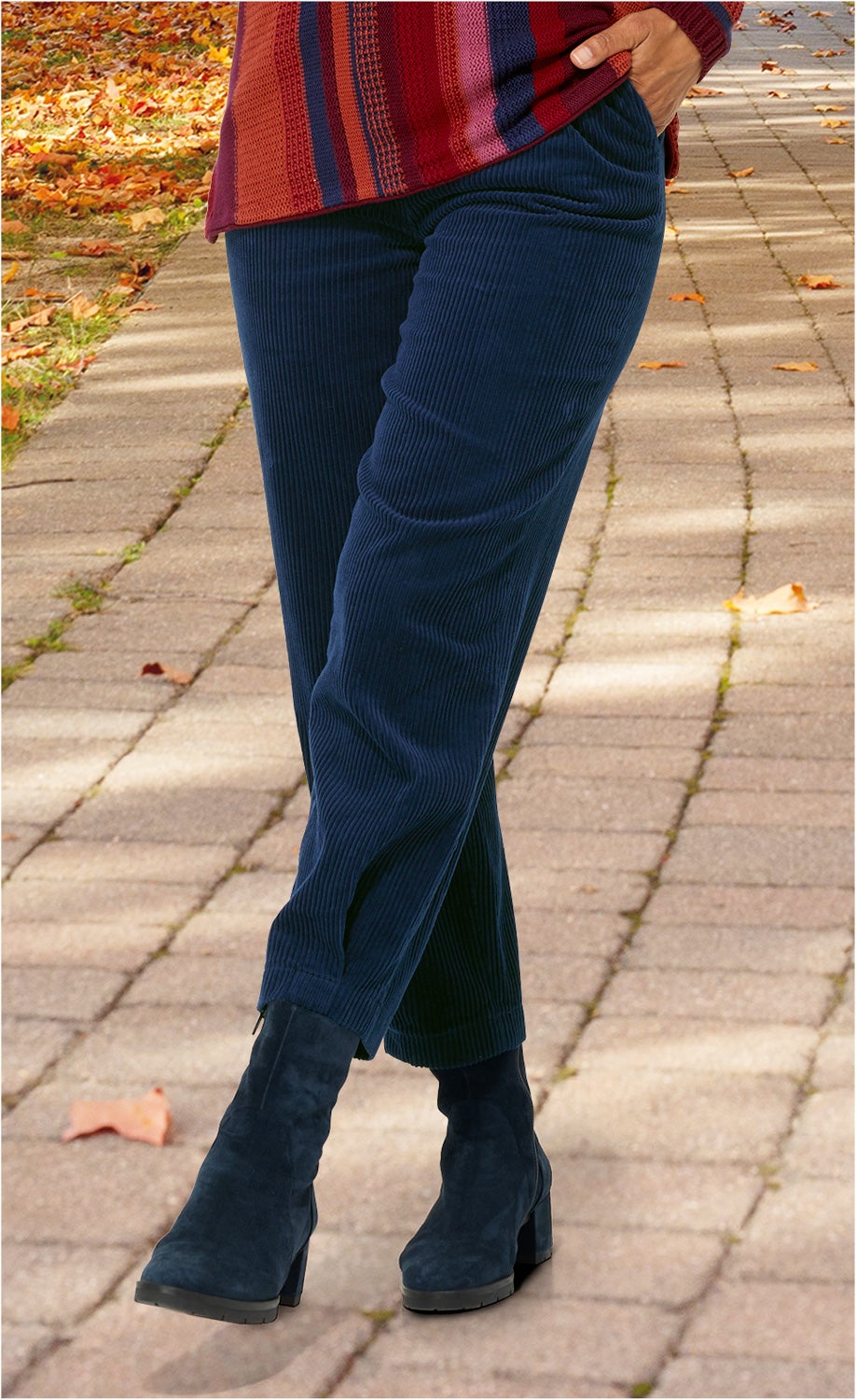 Person trägt dunkelblaue, cordartige Baumwoll-Hose mit engem Bein, dazu dunkelblaue Stiefeletten mit Blockabsatz, Herbstweg mit buntem Laub im Hintergrund, Hand lässig in Hosentasche