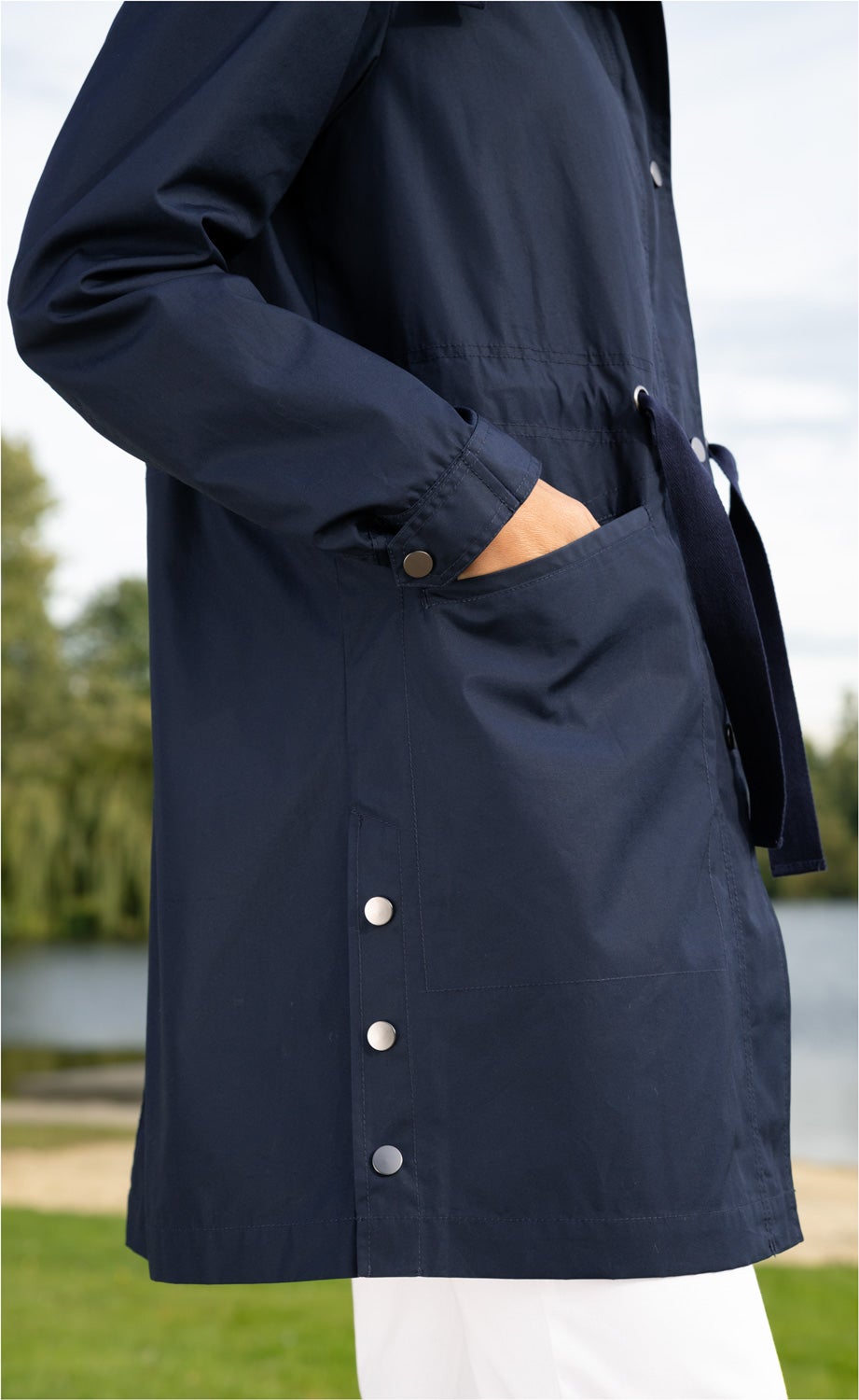 Dunkelblaue Baumwoll-Jacke mit seitlicher Knopfleiste, aufgesetzter Tasche und Kordelzug, Hand in Tasche, weißer Hintergrund, Außenaufnahme mit Wasser und Bäumen im Hintergrund