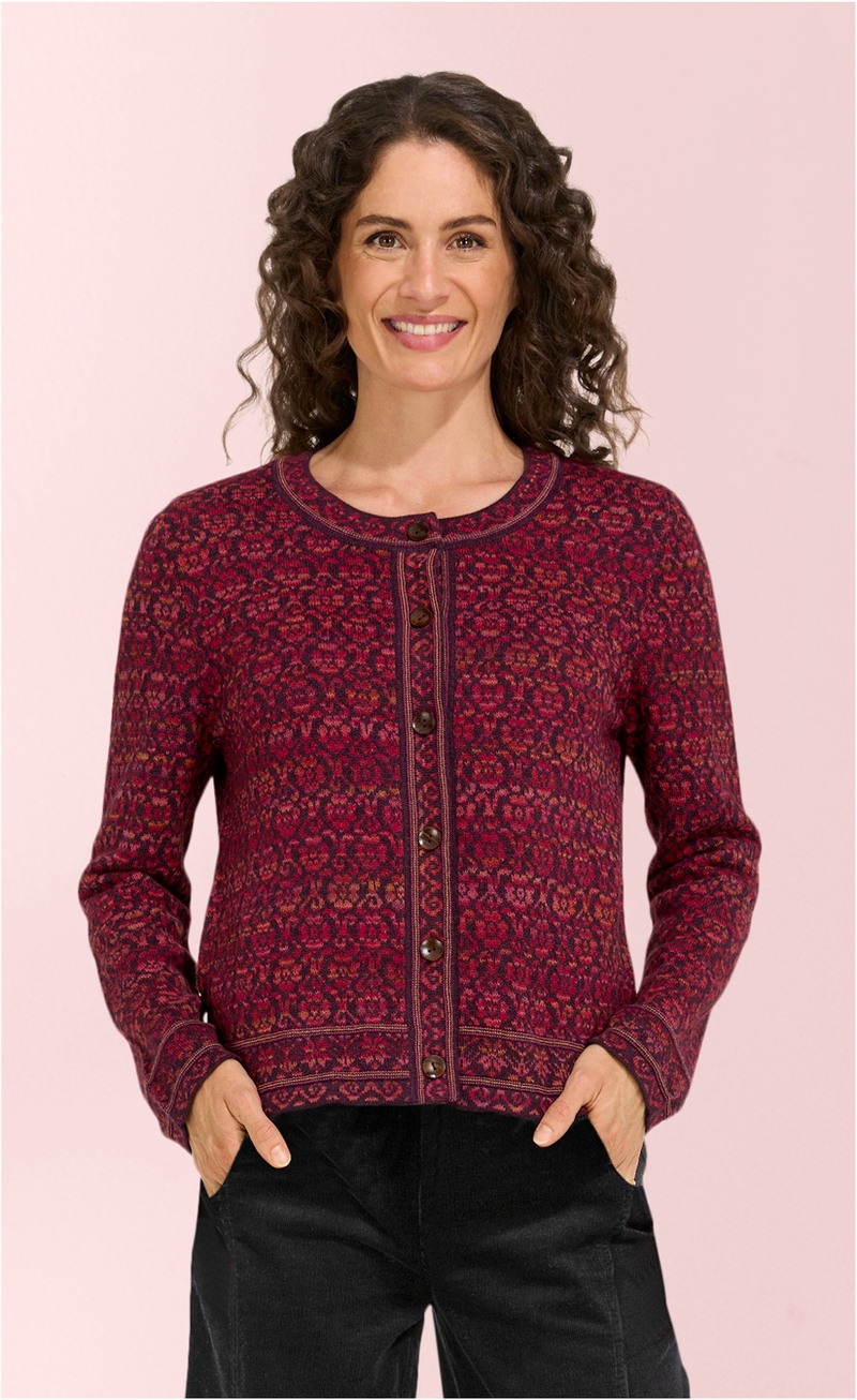 Frau mit gelocktem braunem Haar trägt eine rot-gemusterte Alpaka-Strickjacke mit Rundhalsausschnitt und Knopfleiste, schwarze Hose, Hände in den Hosentaschen, vor hellem rosa Hintergrund.