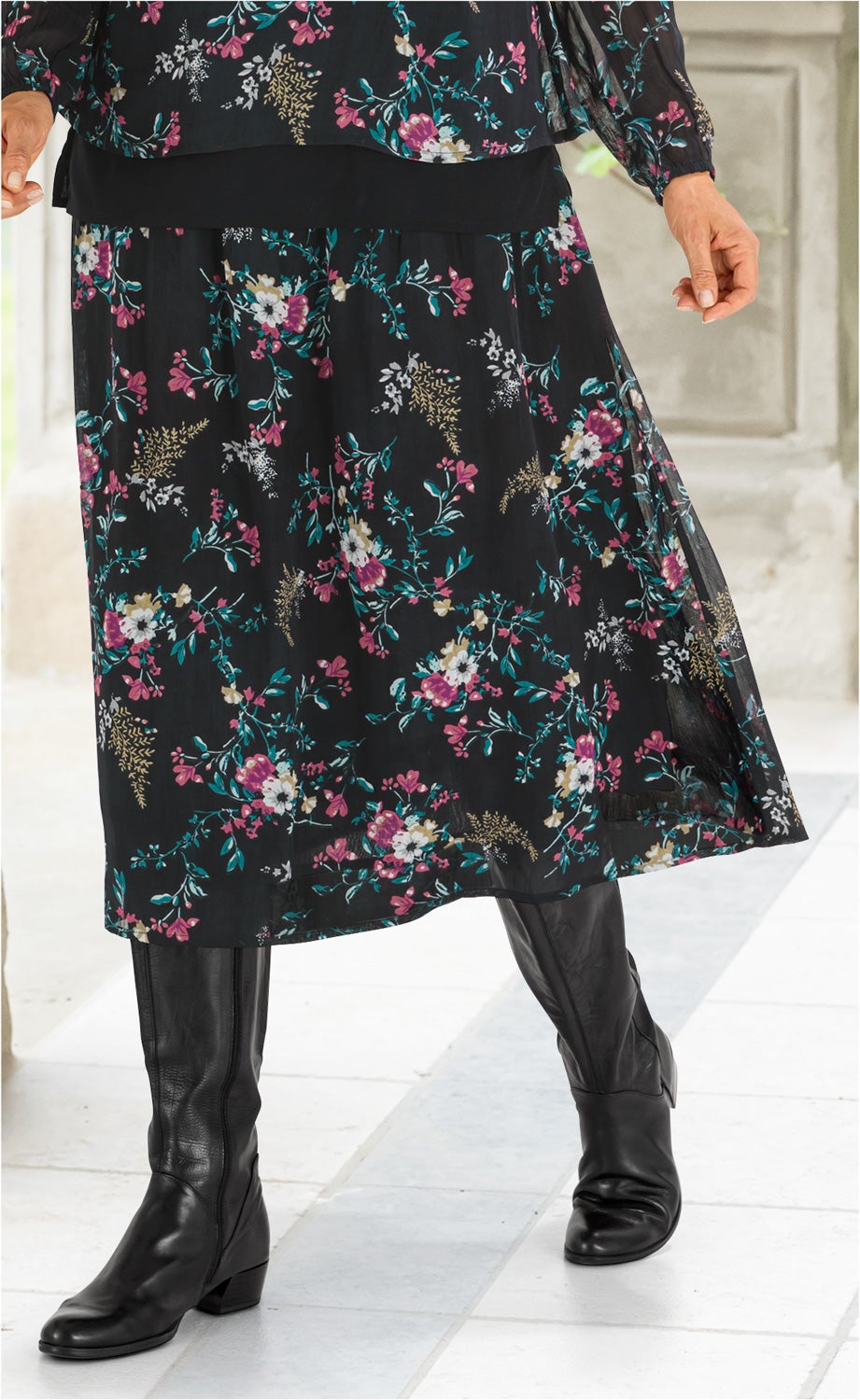 Schwarzer Viskose-Rock mit buntem Blumenmuster, knieumspielend, kombiniert mit schwarzer Transparenzbluse und schwarzen Lederstiefeln.