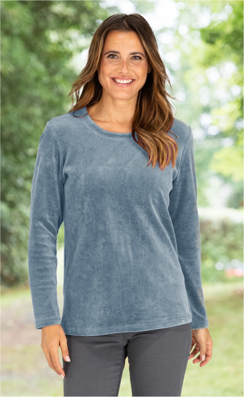 lächelnde Frau mit langen braunen Haaren, trägt hellblaues langärmeliges Shirt und dunkle Hose, unscharfer grüner Hintergrund mit Bäumen und Lichtpunkten