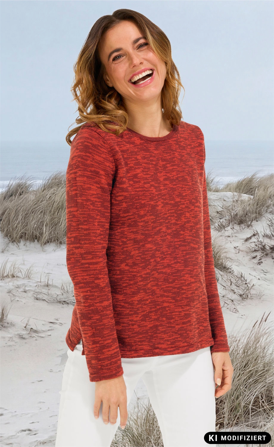 Frau mit lockigen braunen Haaren trägt roten gemusterten Baumwoll-Pullover und weiße Hose, lächelt, steht am Strand mit Sand und Dünen im Hintergrund.