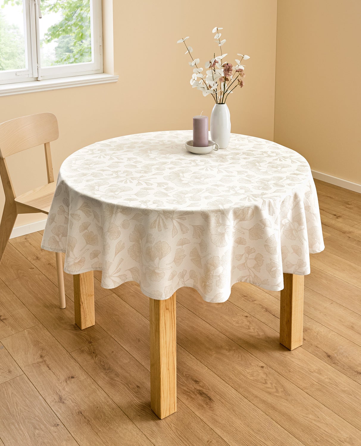 Runder Holztisch mit cremefarbenem Baumwoll-Tischdeck130x180 mit floralem Muster, darüber weiße Vase mit zarten weißen und rosa Trockenblumen und lila Kerze auf hellem Untersetzer, helle Wand und Fenster, einfacher Holzstuhl