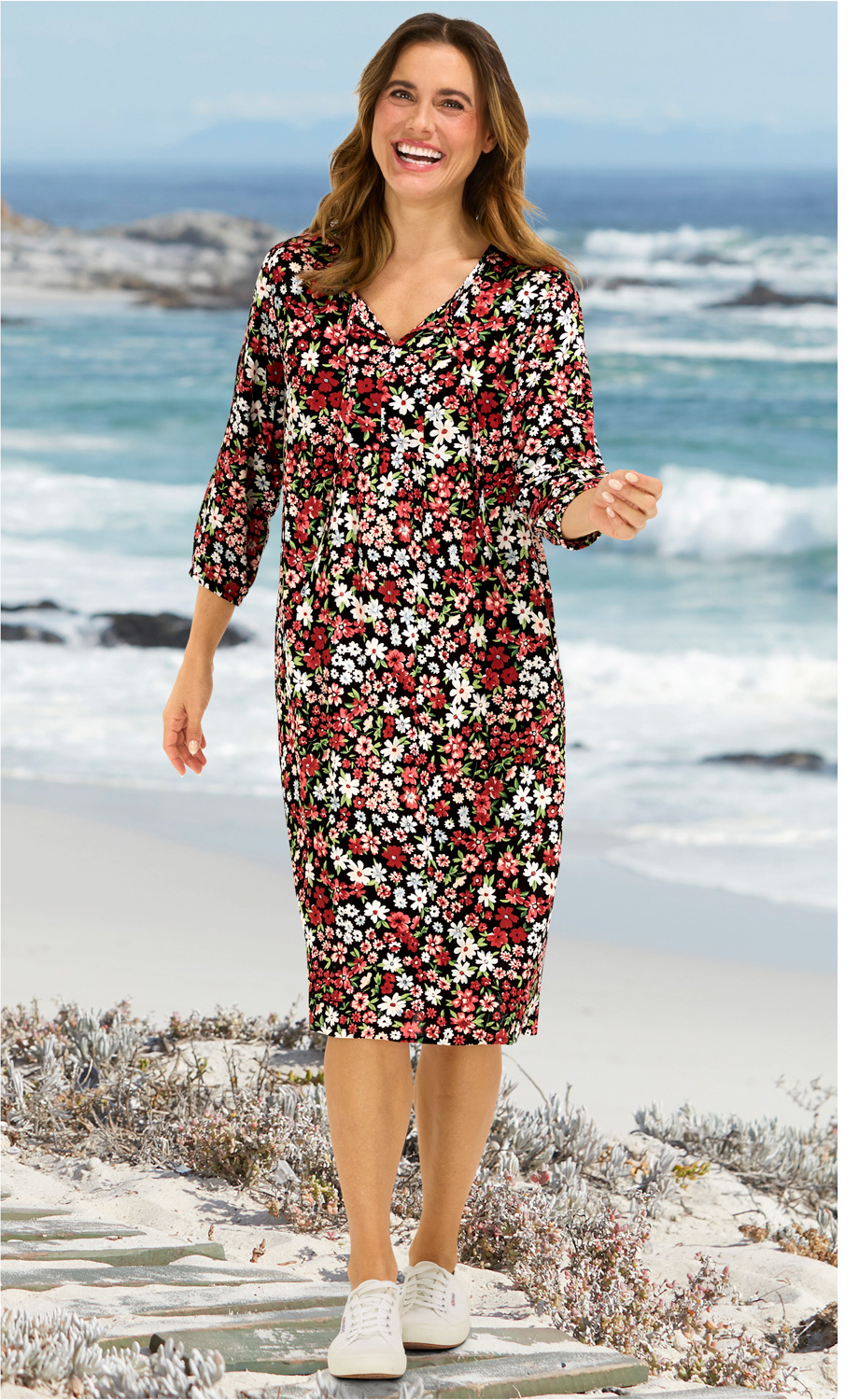 Frau trägt knielanges Viskose-Kleid mit buntem Blumenmuster, V-Ausschnitt und 3/4-Ärmeln, weiße Sneaker, steht am Strand mit Meer und Felsen im Hintergrund, lächelt freundlich