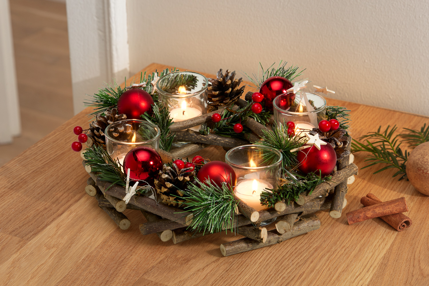 Adventskranz aus kleinen Holzästen, dekoriert mit Tannenzweigen, roten Christbaumkugeln, Zapfen, roten Beeren und vier brennenden Teelichtern in transparenten Glasbehältern, auf Holztisch mit Zimtstangen und Tannenzweig.