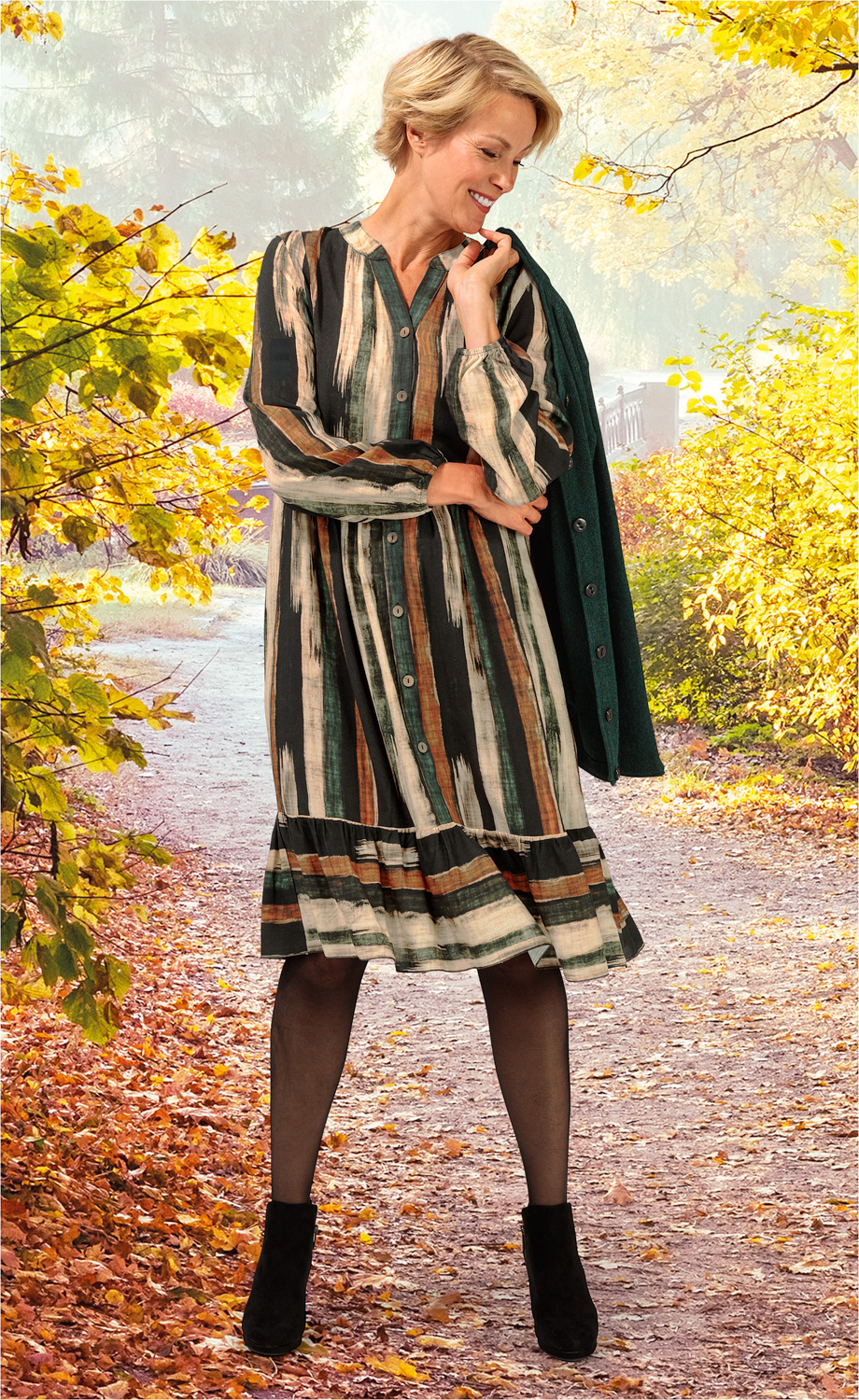 Frau trägt gestreiftes Viskose-Kleid in Grün-, Braun- und Beigetönen, schwarze Strumpfhose und schwarze Stiefeletten, hält grünen Mantel über der Schulter, steht auf herbstlichem Waldweg mit gelb und orange gefärbtem Laub