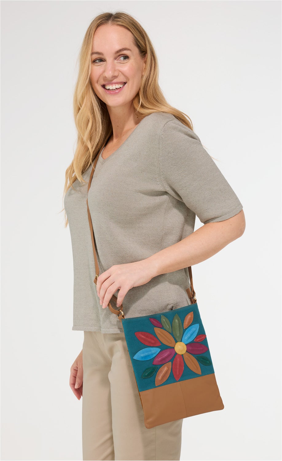 Frau trägt beige Schultertasche mit buntem Blumenmuster aus Leder und Stoff, hellgrauer Pullover mit halblangen Ärmeln, beigefarbene Hose, lächelt, weißer Hintergrund