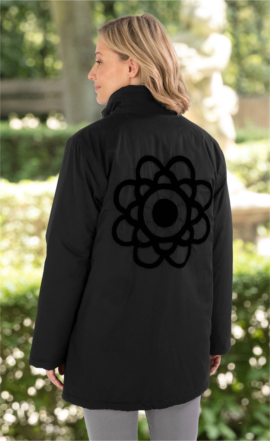 Frau von hinten in schwarzer Baumwoll-Jacke mit Stehkragen und dekorativem, schwarzem Blumenmuster auf dem Rücken, in hellem Outdoor-Garten mit unscharfem Hintergrund aus Bäumen und Skulpturen.