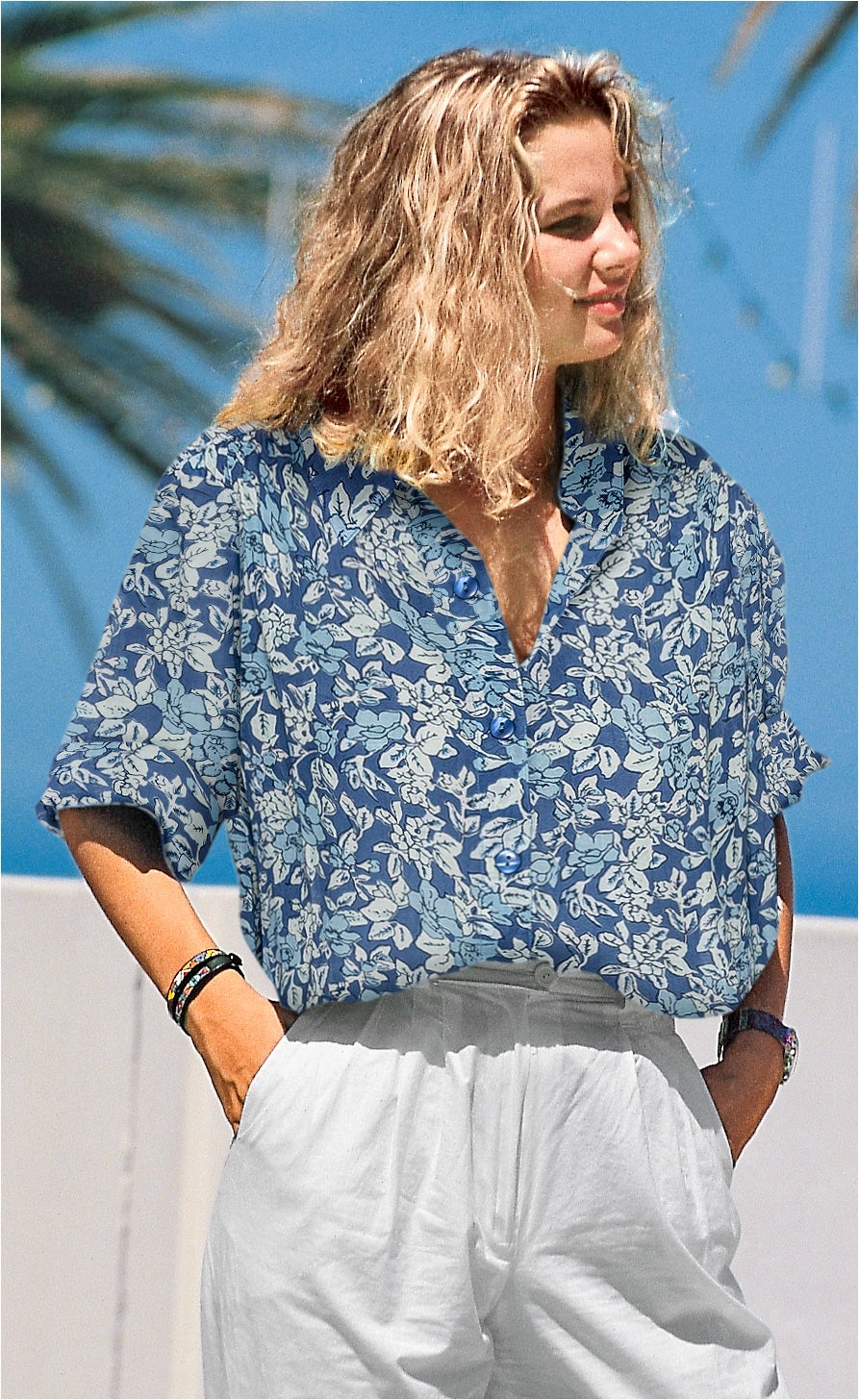 Frau mit halblangen welligen blonden Haaren trägt blau-weiße Viskose-Mix-Bluse mit floralem Muster, weißer Hose, Armbändern und Armbanduhr, Sommerhintergrund mit Palmen und blauem Himmel
