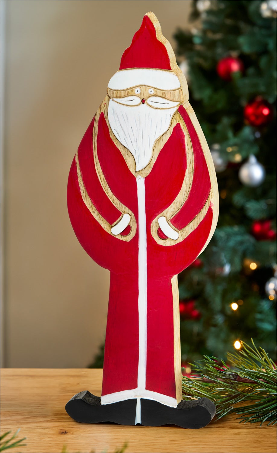Holzfigur eines Weihnachtsmanns in rotem Mantel und Mütze, mit weißem Bart, stehend auf schwarzem Sockel, im Hintergrund unscharfer Weihnachtsbaum mit roten und silbernen Kugeln, auf Holzuntergrund mit Tannenzweig.
