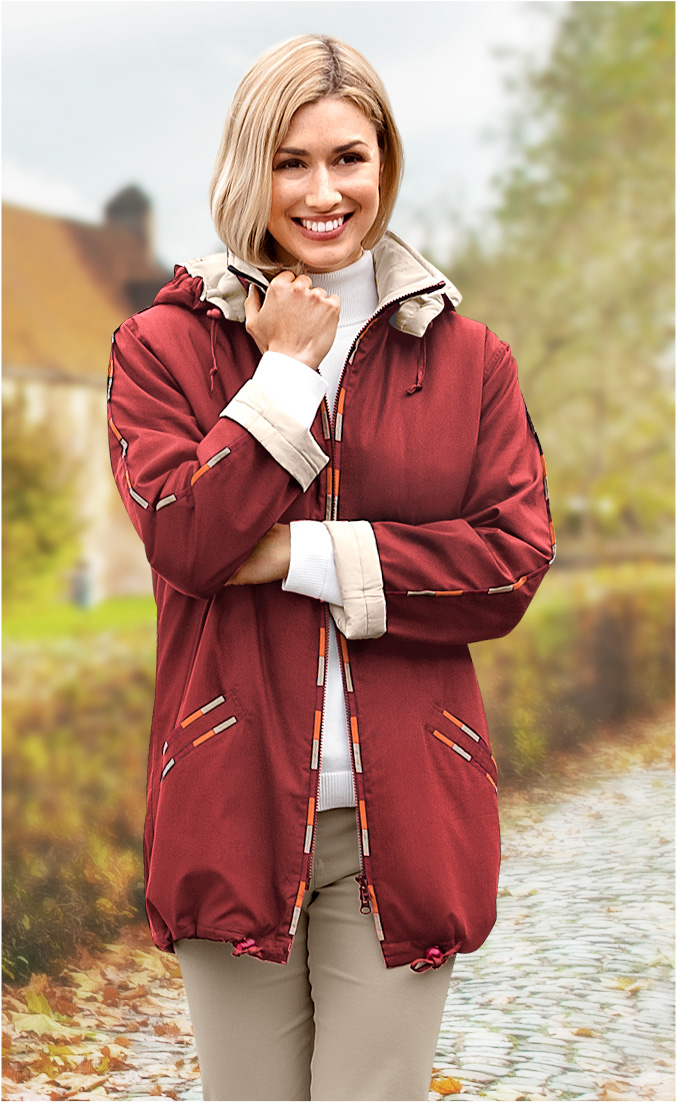 Frau trägt rote Baumwoll-Jacke mit beigen Akzenten an Kragen und Ärmelbündchen, weißer Rollkragenpullover, beige Hose, steht lächelnd auf gepflastertem Weg mit Laub im Hintergrund