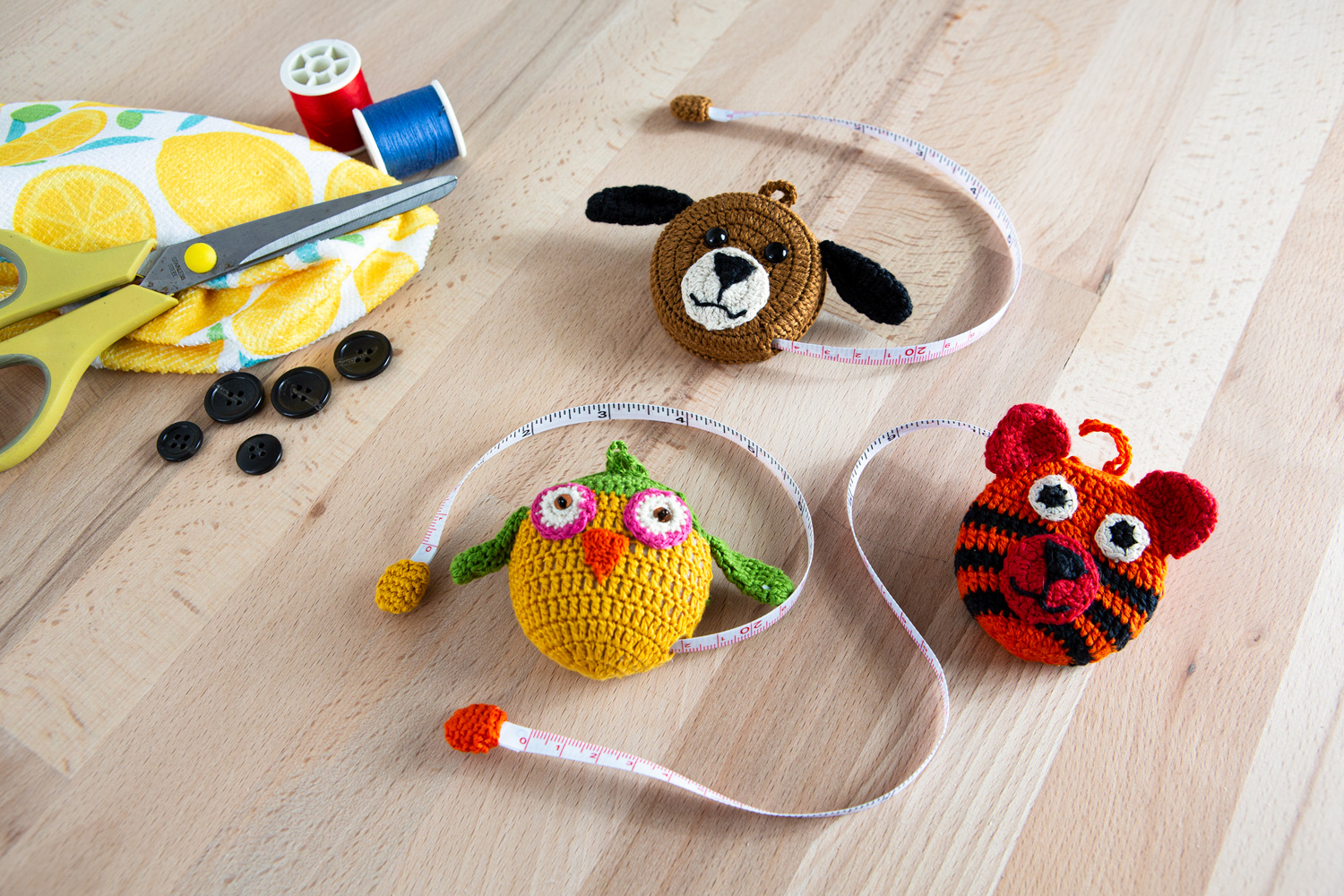 Maßband in Form einer gehäkelten Eule mit grünem Kopf und gelbem Körper auf hellem Holztisch, daneben bunte gehäkelte Tier-Maßbänder, gelbe Schere, Nähgarnspulen und schwarze Knöpfe