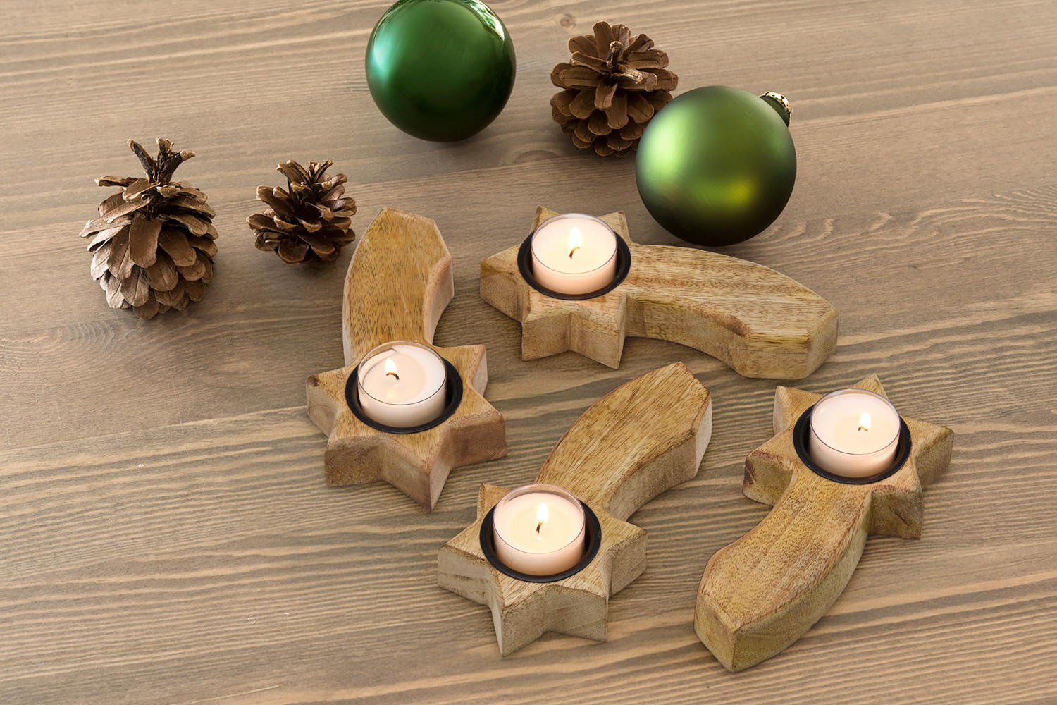 Vier Holz-Teelichthalter in Stern- und Kometenform mit brennenden Teelichtern auf Holztisch, dekoriert mit vier braunen Zapfen und zwei grünen Weihnachtskugeln