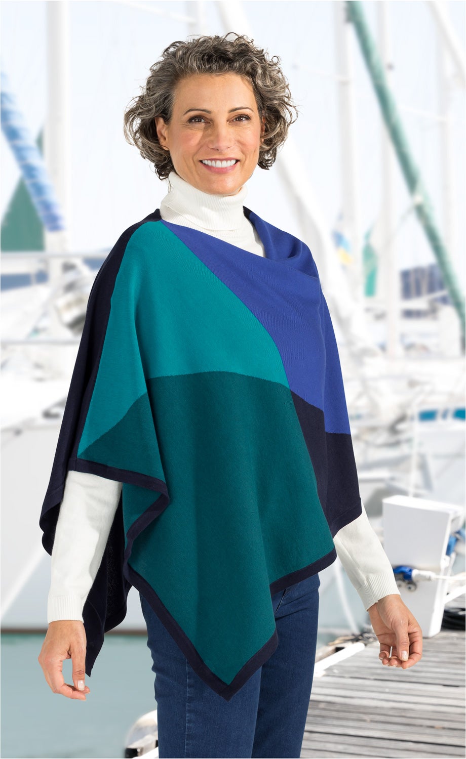 Frau mit kurzen lockigen Haaren trägt farblich gestalteten Baumwoll-Mix-Poncho in Blau- und Grüntönen über weißem Rollkragenpullover, stehender Pose an einem Yachthafen auf Holzsteg