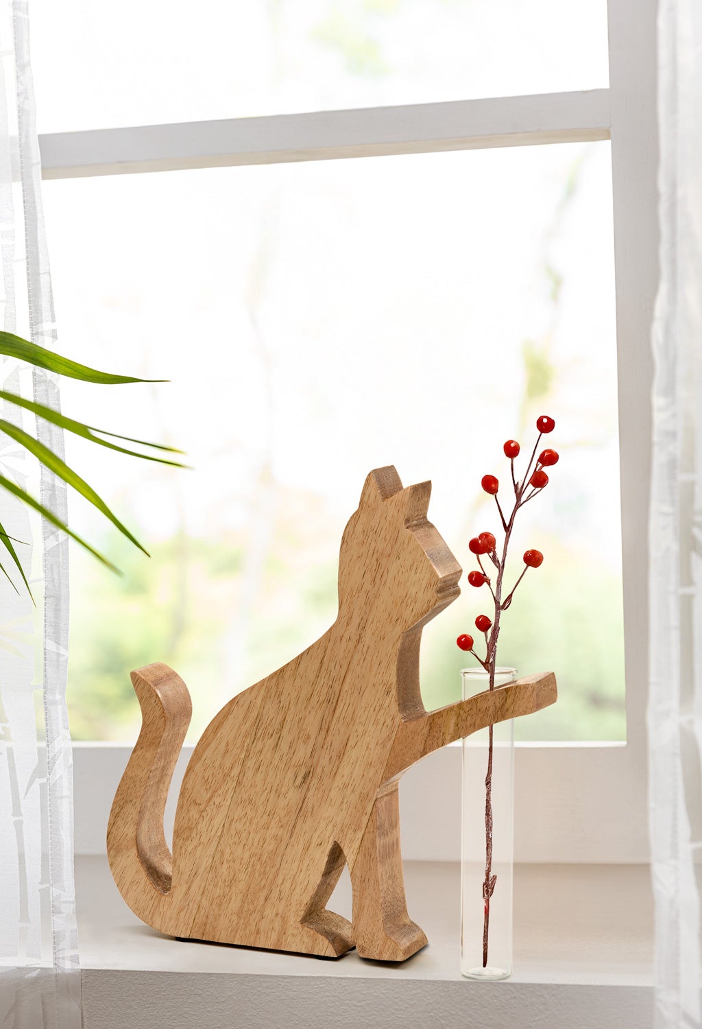 Holzfigur einer Katze mit einer Pfote auf einer kleinen Glasvase, die einen Zweig mit roten Beeren enthält, auf einem weißen Fensterbrett vor hellem Hintergrund mit grünen Pflanzen und weißen Vorhängen