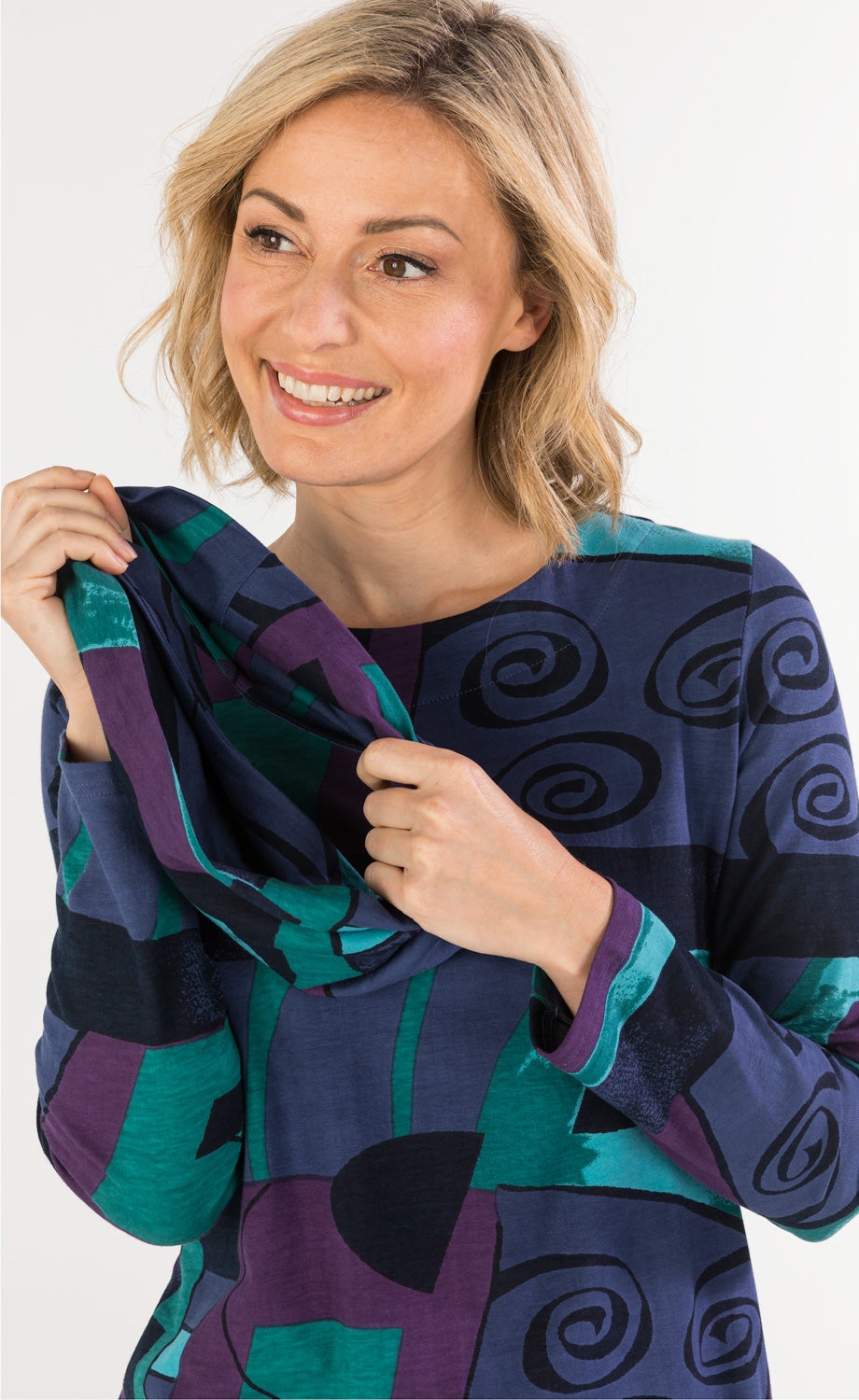 Frau mit blondem Bob-Look lächelt, trägt buntes Baumwoll-Mix-Shirt mit abstraktem Muster in Blau, Lila, Grün, hält den Kragen des langärmligen Shirts mit beiden Händen gespannt.