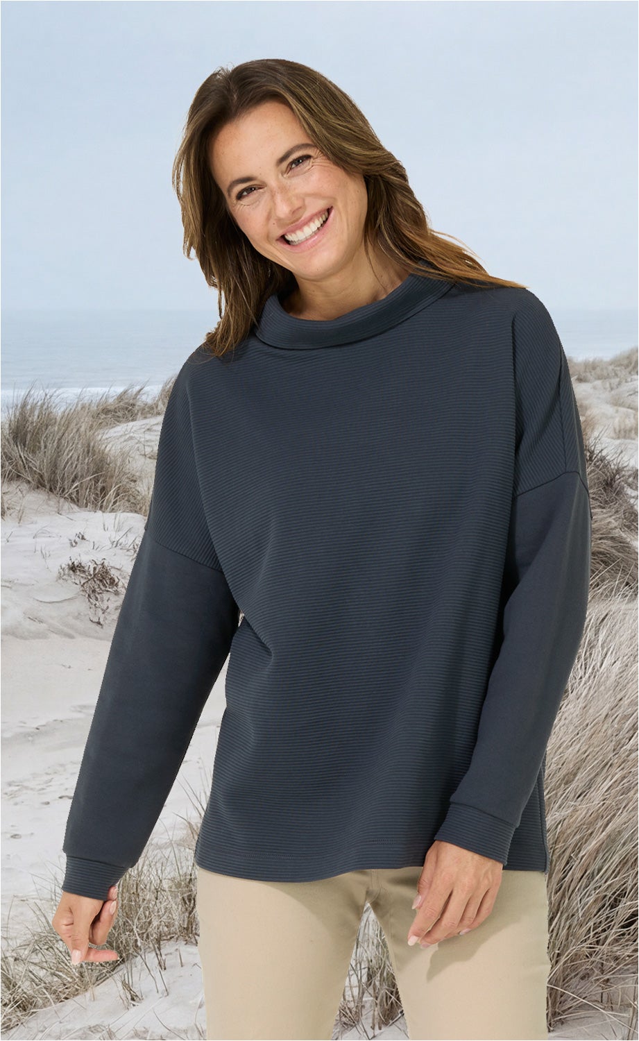 Frau mit braunem Haar trägt langärmliges dunkelblaues Baumwoll-Shirt mit hohem Kragen, steht lächelnd am Strand mit Dünen im Hintergrund, beige Hose, natürliche Pose