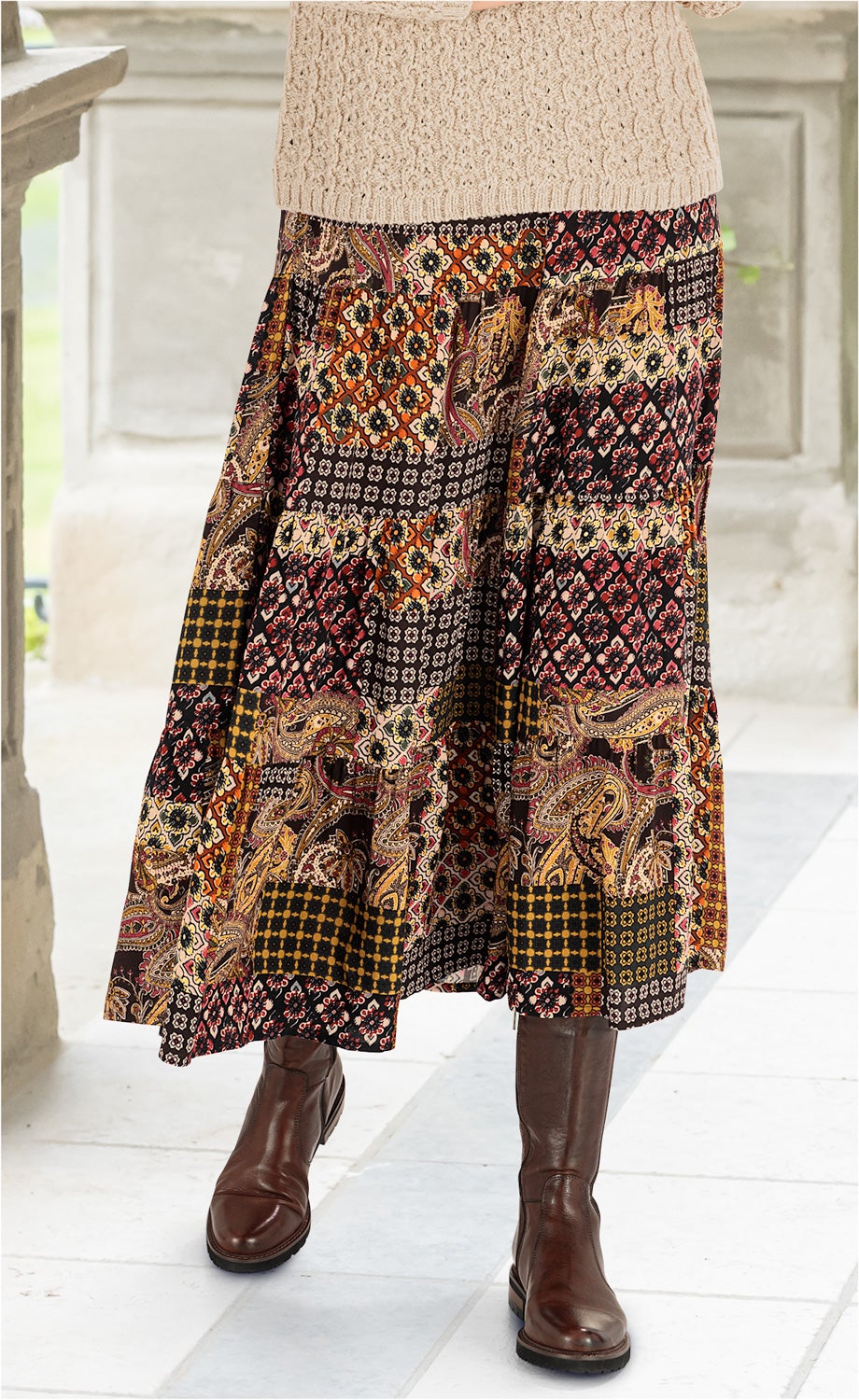 Viskose-Rock mit buntem Patchwork-Muster in Rot-, Gelb- und Schwarztönen, kombiniert mit braunen Lederstiefeln und beige-grauem Grobstrickpullover, aufgenommen auf hell gebfließtetem Boden vor hellem Hintergrund