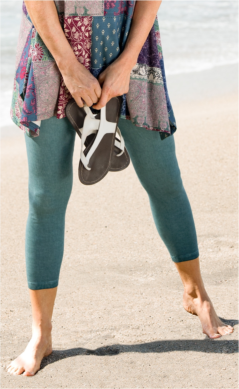 Beinbereich einer Person am Strand, trägt blaugrüne Baumwoll-Mix-Leggings bis knapp über die Knöchel, hält weiße Sandalen in den Händen, bunt gemusterte Tunika, barfuß im Sand stehend, Meeresufer im Hintergrund