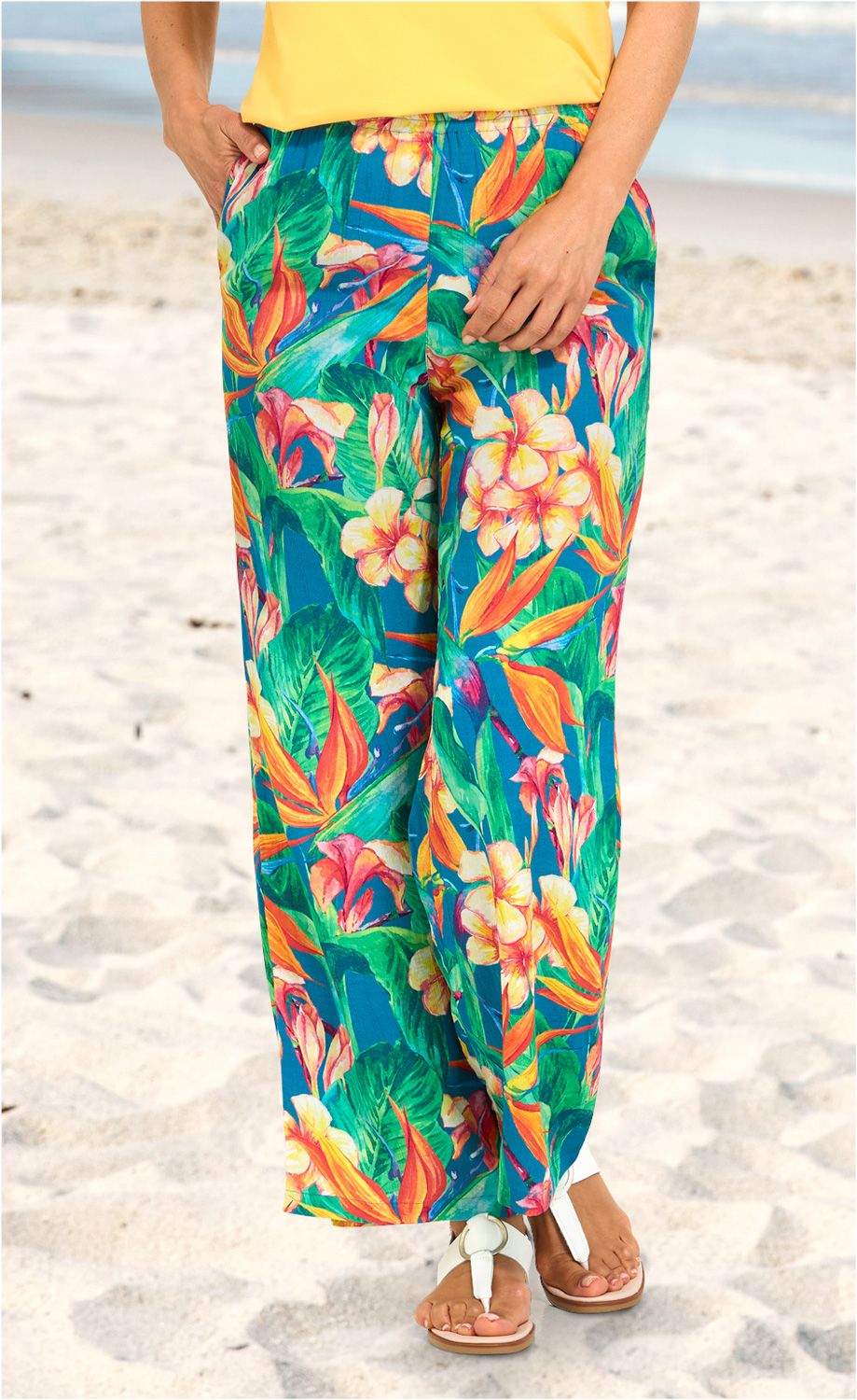 Viskose-Hose mit weitem Bein, lebhaftem tropischem Blumen- und Blätterprint in Grün, Orange, Gelb und Pink, kombiniert mit gelbem Oberteil und weißen Sandalen, aufgenommen am Strand auf Sandboden