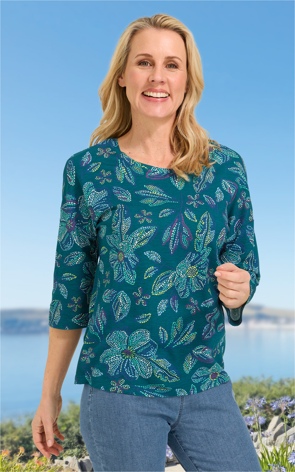 Frau mit blau-grünem Viskose-Shirt mit floralem Muster und 3/4-Ärmeln, lächelnd, vor sommerlichem Hintergrund mit blauem Himmel und Wasser, trägt blaue Jeans