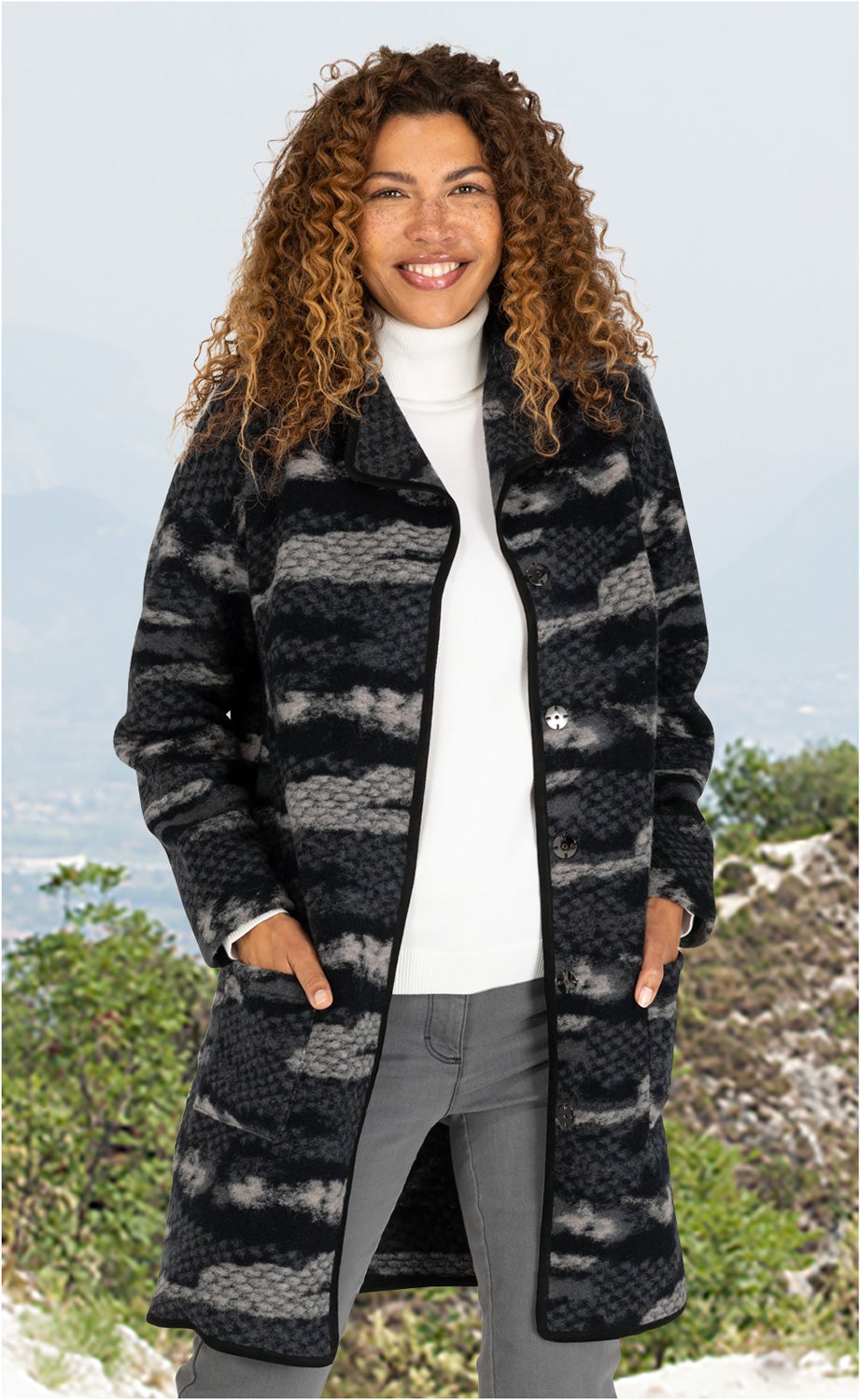 Frau trägt Woll-Mix-Mantel mit dunklem Muster, weißem Rollkragenpullover und grauer Hose vor Berglandschaft, Hände in Manteltaschen, lockige Haare, lächelnd