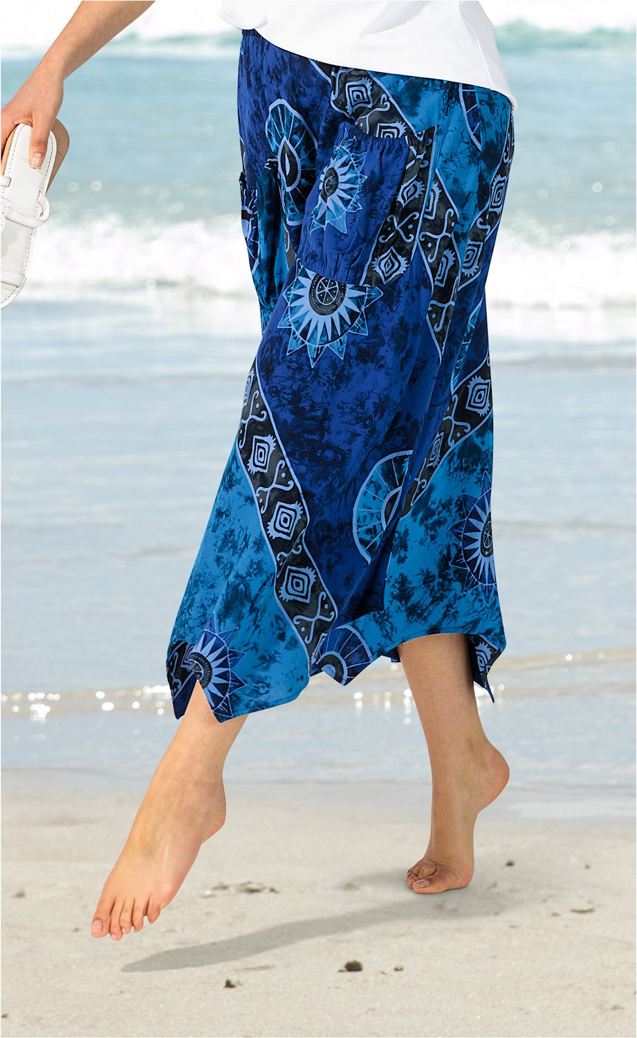 Frau am Strand mit blau gemustertem Viskose-Rock und weißen Sandalen in der Hand, barfuß im Sand, Hintergrund Meer und Wellen, lebendige Farben und sommerliches Strandmotiv