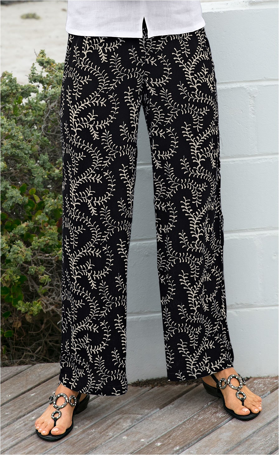 Schwarze Viskose-Hose mit beigem, verzweigtem Blattmuster, weiter Schnitt, getragen mit schwarzen Sandalen mit großen, silbernen Ringen, teilweise sichtbares weißes Oberteil, Holzboden und weiße Mauer im Hintergrund