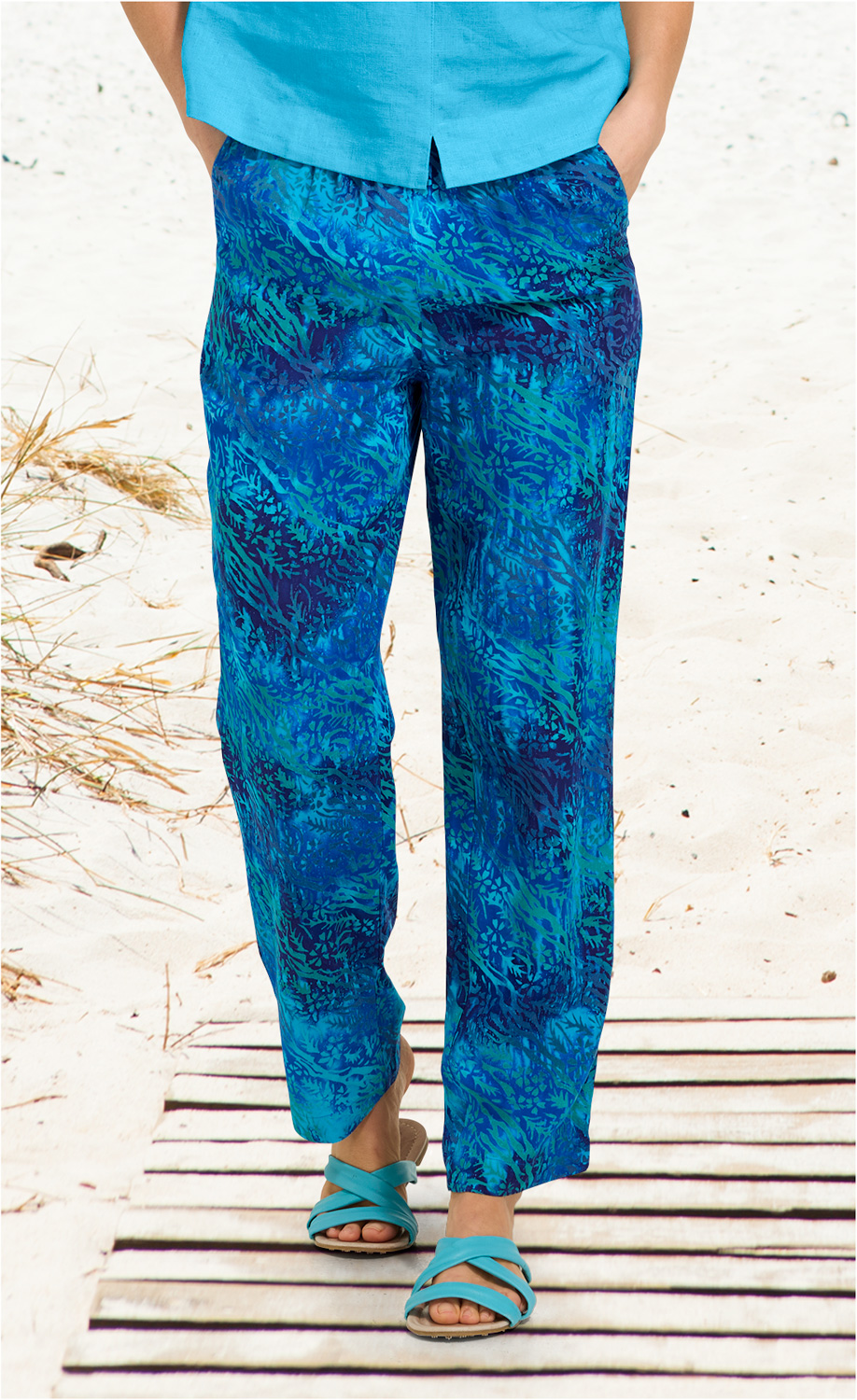 Blaue Viskose-Hose mit abstraktem Muster in verschiedenen Blautönen, weit geschnitten, getragen mit türkisfarbenen Sandalen, Strandhintergrund, Hände in den Hosentaschen, türkisfarbenes Oberteil teilweise sichtbar