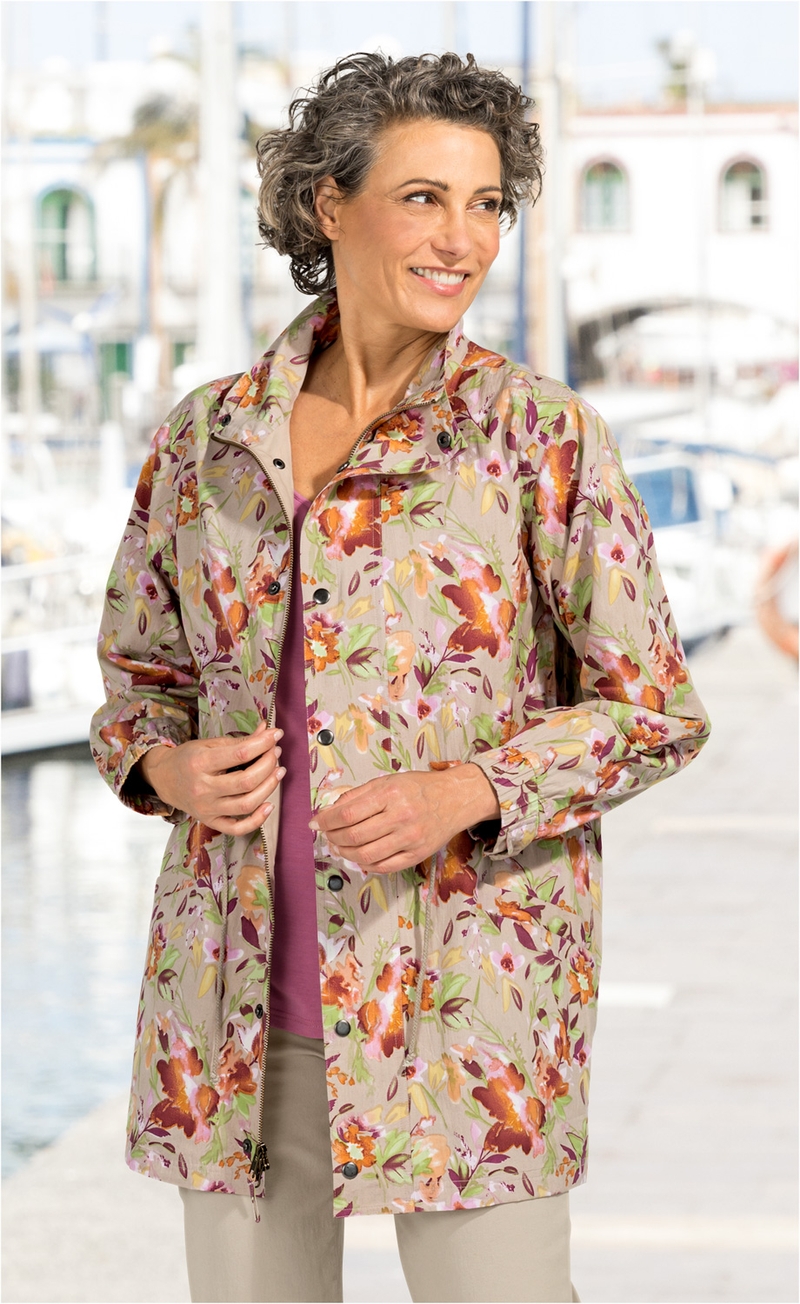 Frau trägt leichte Baumwoll-Jacke mit buntem Blumenmuster in Beige, Orange, Grün und Pink, kombiniert mit beiger Hose und rosafarbenem Oberteil, steht vor unscharfem Hafen mit Booten im Hintergrund