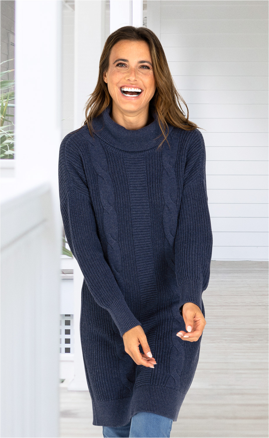 lächelnde Frau trägt navyblaues, langärmeliges Baumwoll-Mix-Kleid mit Zopfmuster und Rollkragen, stehender Raum mit weißer Holzverkleidung im Hintergrund
