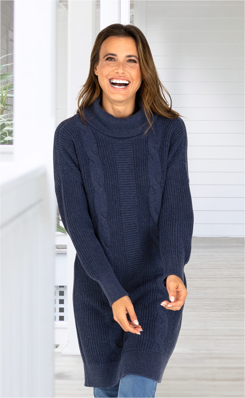 lächelnde Frau trägt navyblaues, langärmeliges Baumwoll-Mix-Kleid mit Zopfmuster und Rollkragen, stehender Raum mit weißer Holzverkleidung im Hintergrund