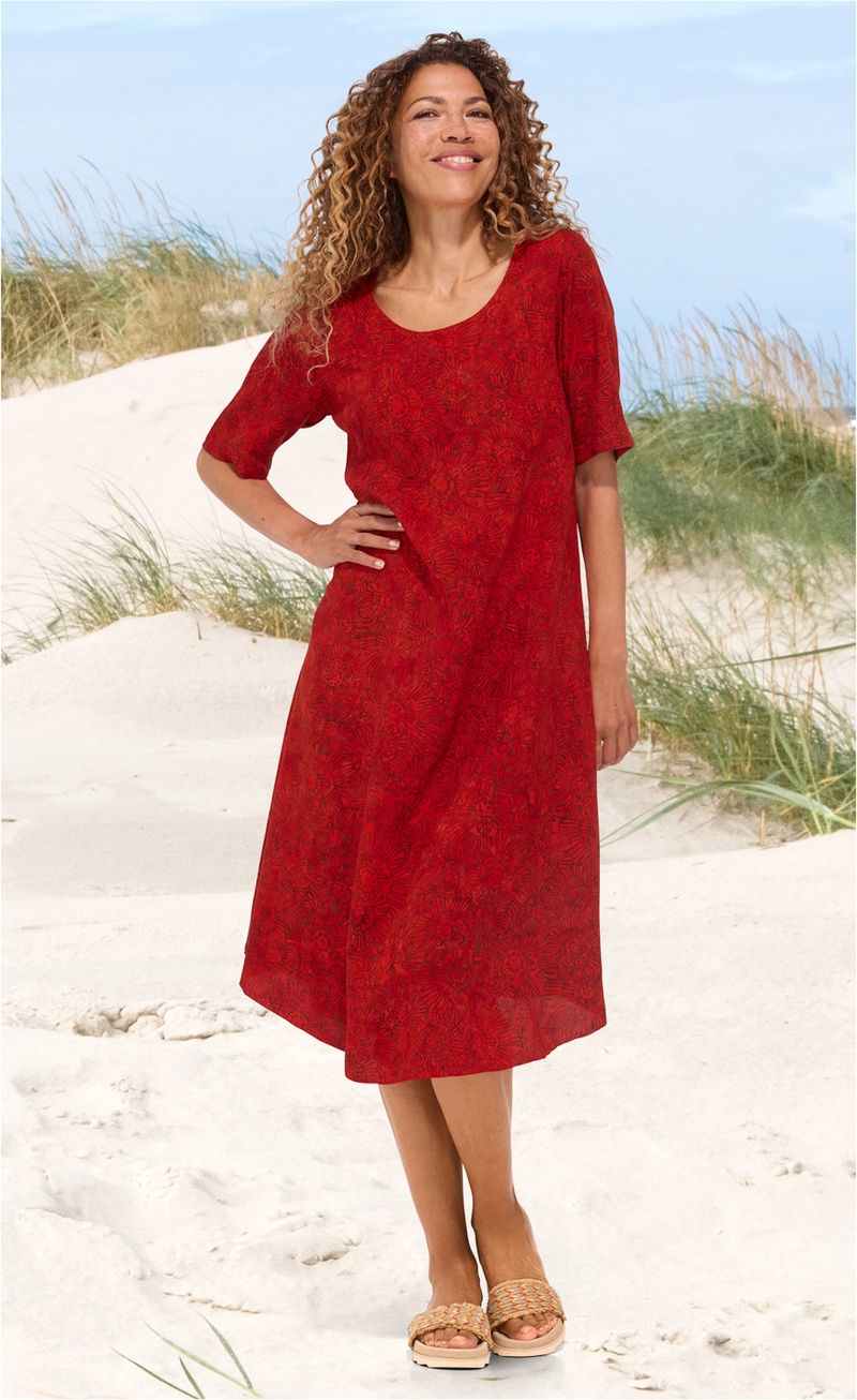 Frau am Strand trägt rotes Viskose-Kleid mit floralem Muster, halblangen Ärmeln und rundem Ausschnitt, dazu geflochtene Sandalen, lockige Haare, lächelnd, natürliche Umgebung mit Sand und Dünen im Hintergrund
