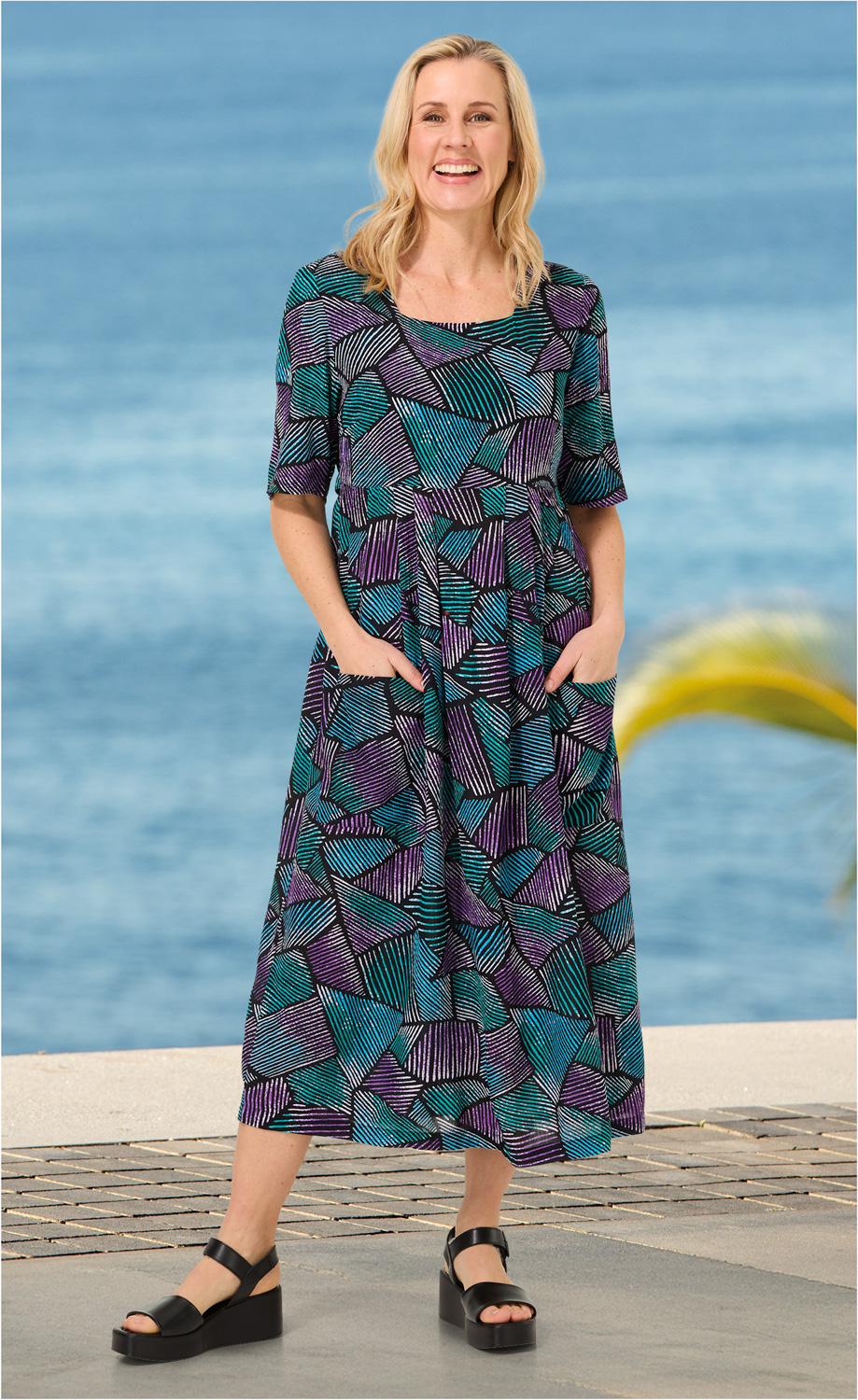 Frau trägt bunt gemustertes Viskose-Kleid mit lila und türkisfarbenen geometrischen Mustern, halblangen Ärmeln und Taschen, dazu schwarze Plateausandalen, Hintergrund Meer und Promenade